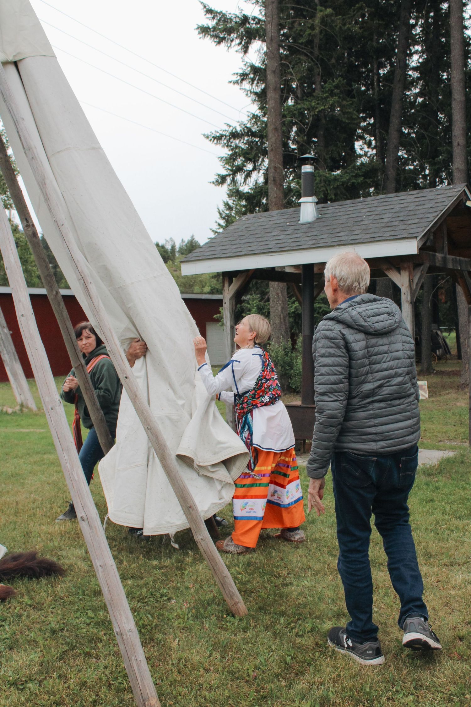 People in traditional Indigenous clothing help assemble a teepee outdoors near a wooden shelter surrounded by tall trees.