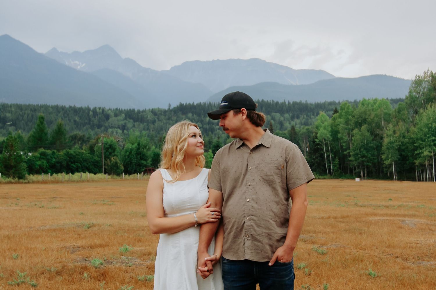 Couple holding hands in a golden field with misty mountains and evergreen forest backdrop on a cloudy day.