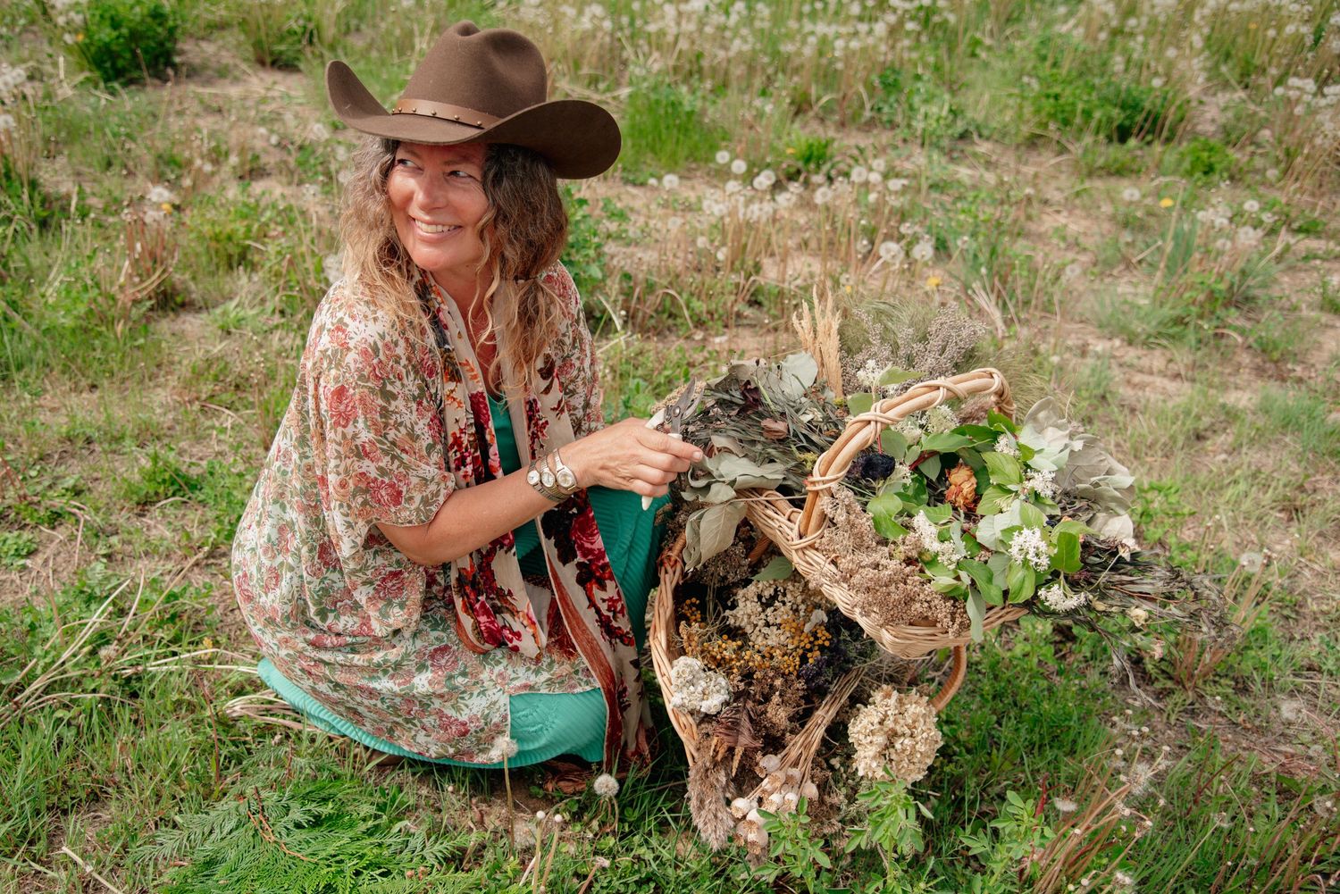 Woman in floral kimono and cowboy hat kneeling in a field with a wicker basket of wildflowers.