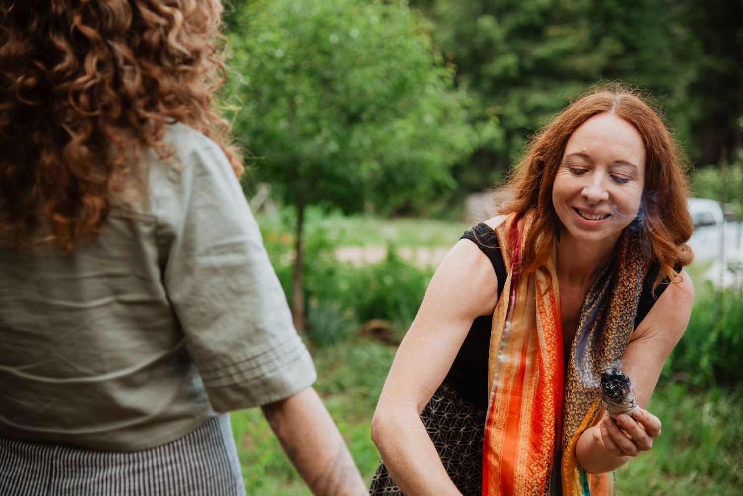 Two red-haired women talking outdoors in a lush green garden, one smiling and holding a small object.