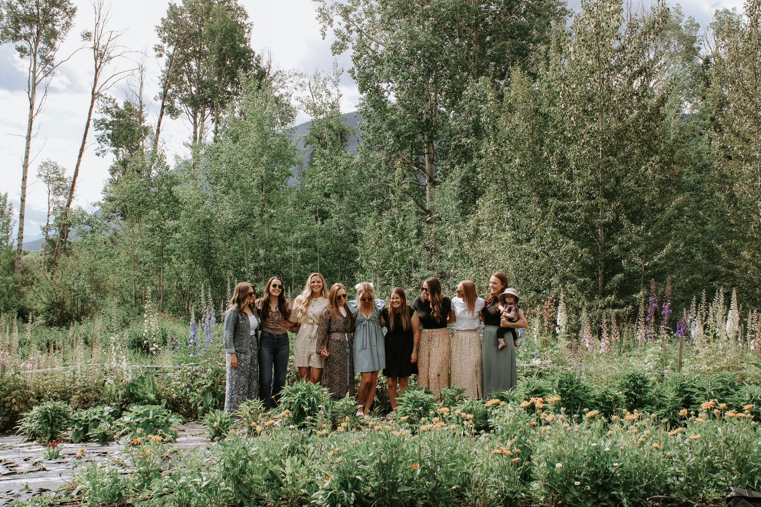 Wedding group photo arranged in a natural outdoor setting with tall trees and wildflowers.