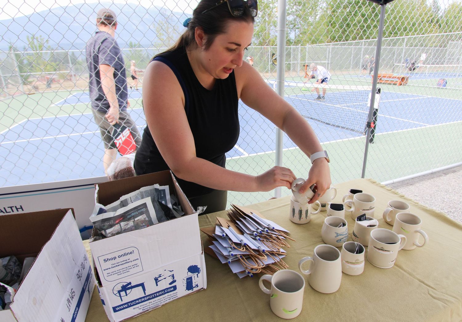 Woman arranging ceramic mugs and brochures at an outdoor vendor table near tennis courts on a sunny day.