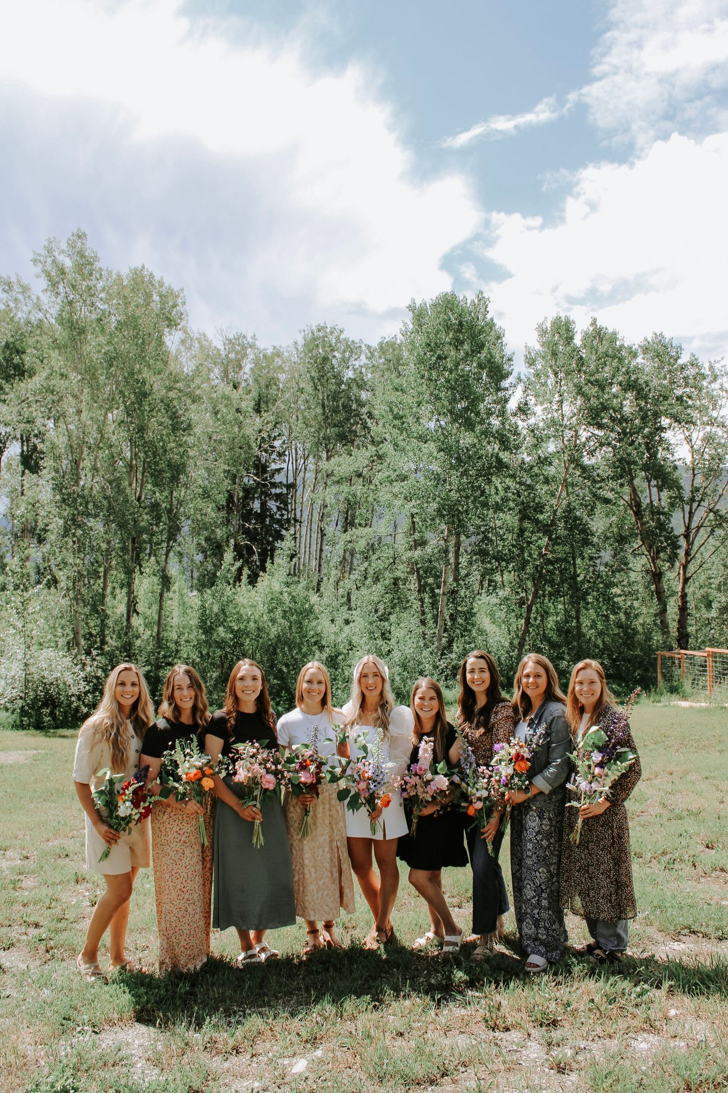 Group of women holding colorful floral bouquets outdoors in a green meadow with tall trees on a sunny day.