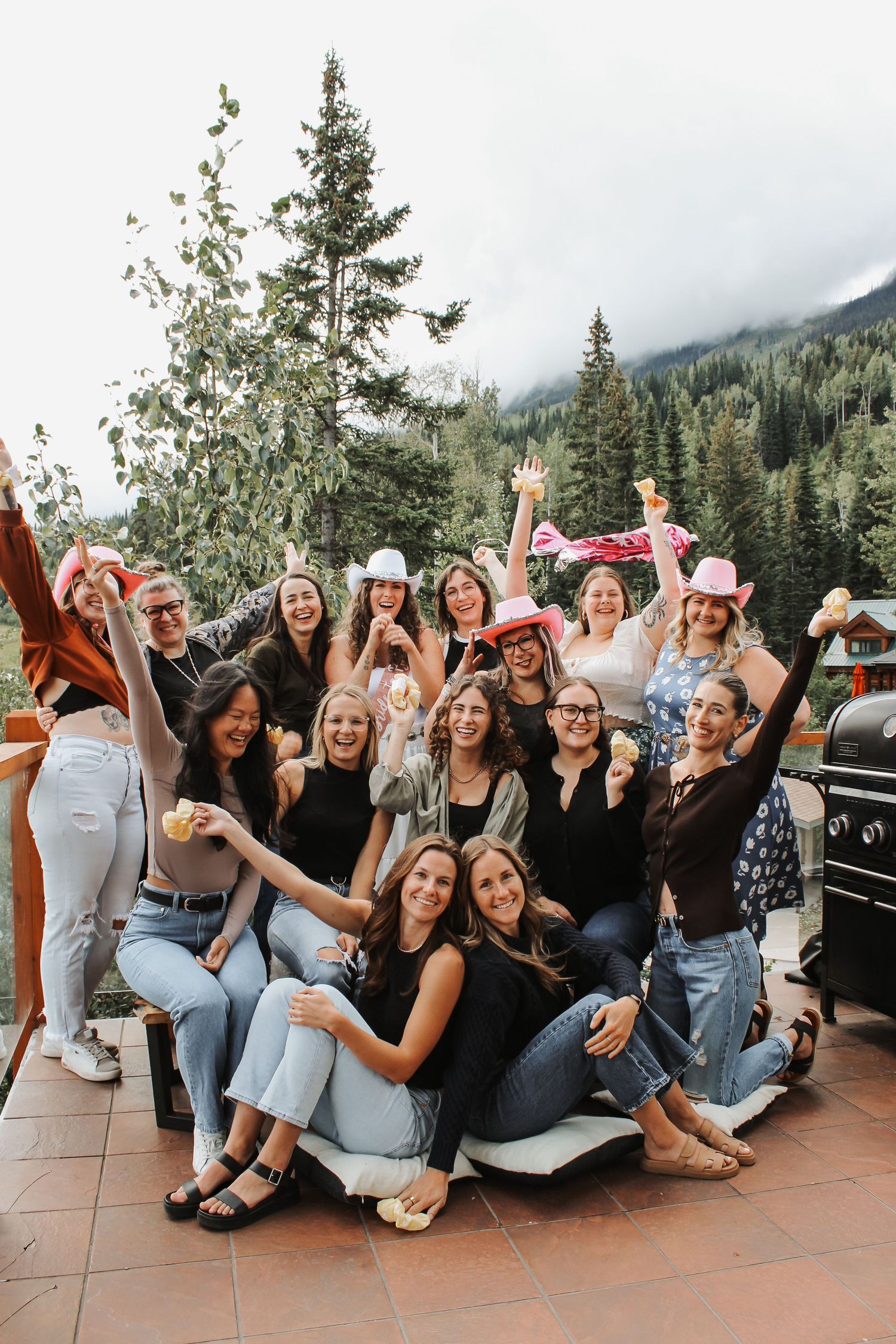 Group of women celebrating a bachelorette party on a mountain deck, wearing pink cowboy hats and raising drinks.