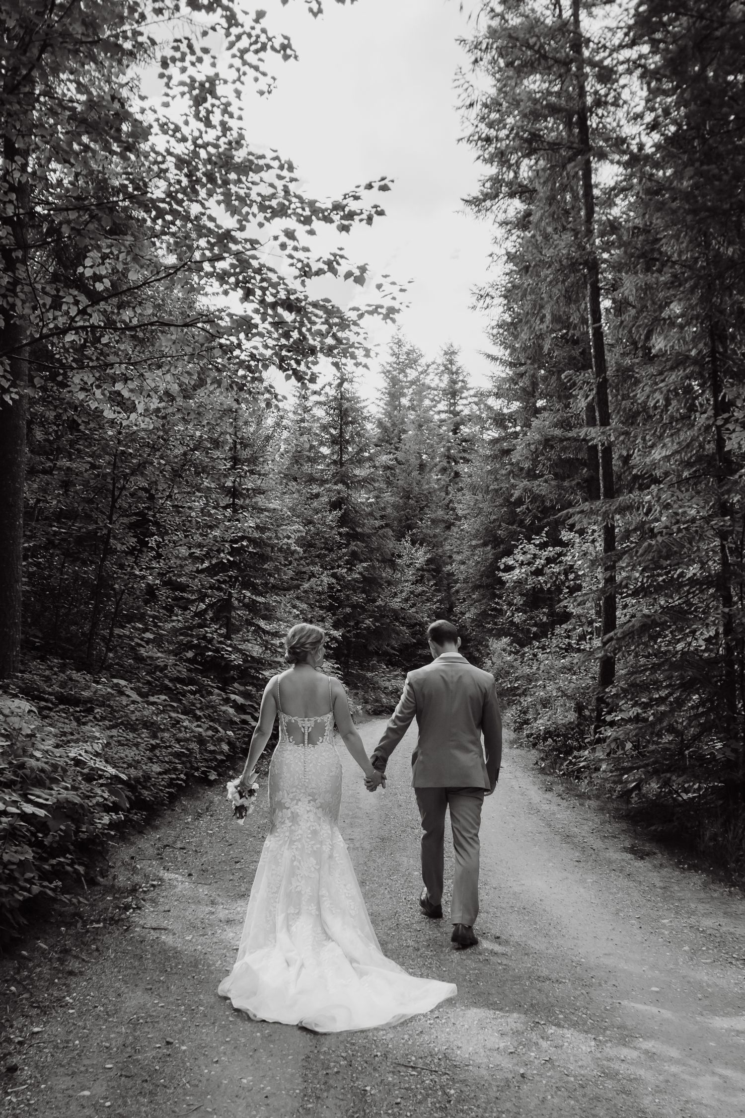 Bride and groom walk hand in hand down a tree-lined forest path in black and white wedding portrait.