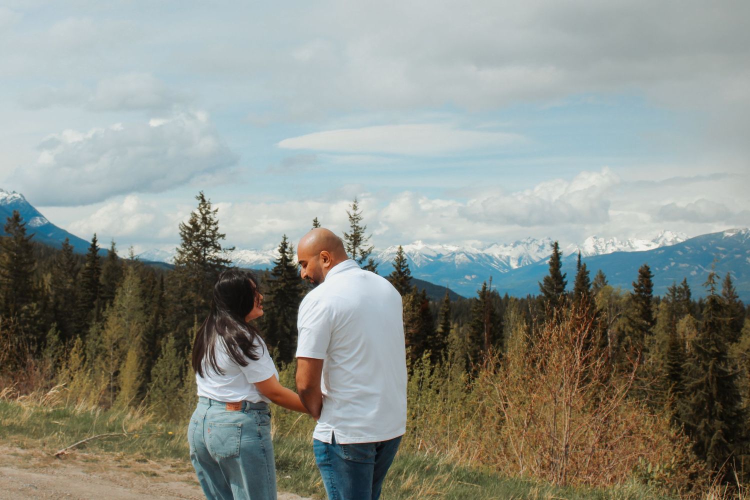 Couple holding hands facing snow-capped mountains and evergreen forest under cloudy sky in scenic outdoor setting.