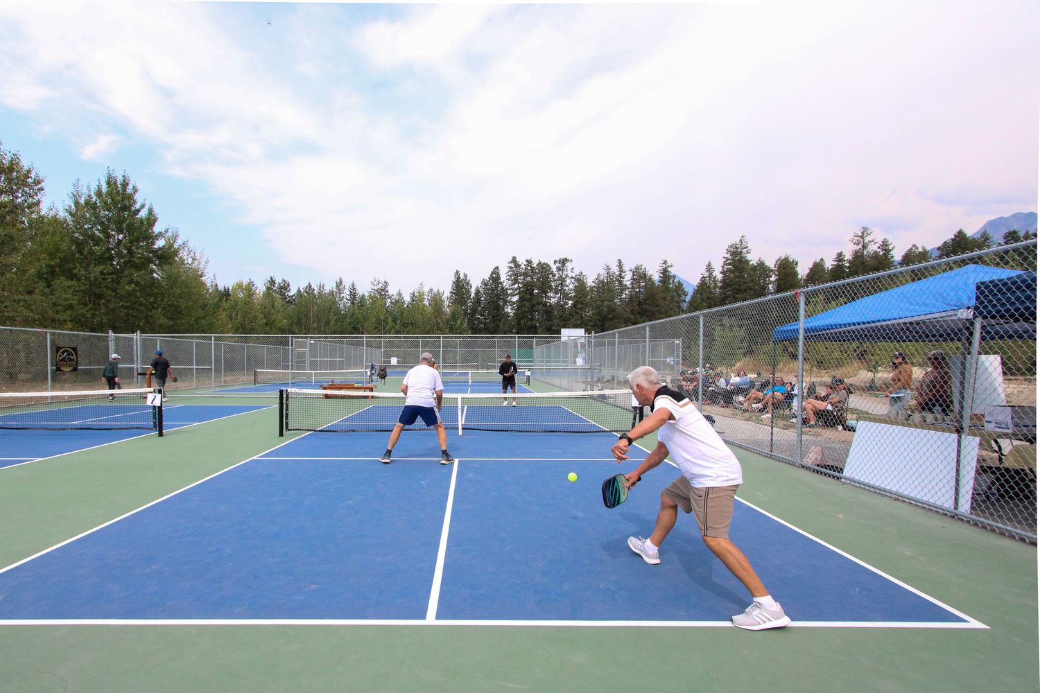 People playing pickleball on a blue outdoor court surrounded by trees and fencing on a partly cloudy day.