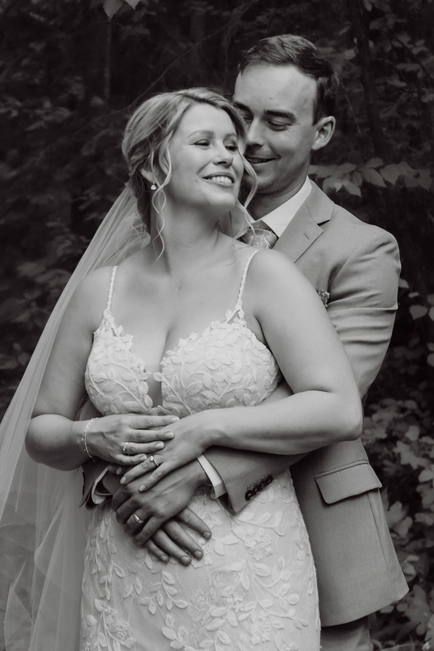 Black and white portrait of smiling bride and groom embracing in forest setting.