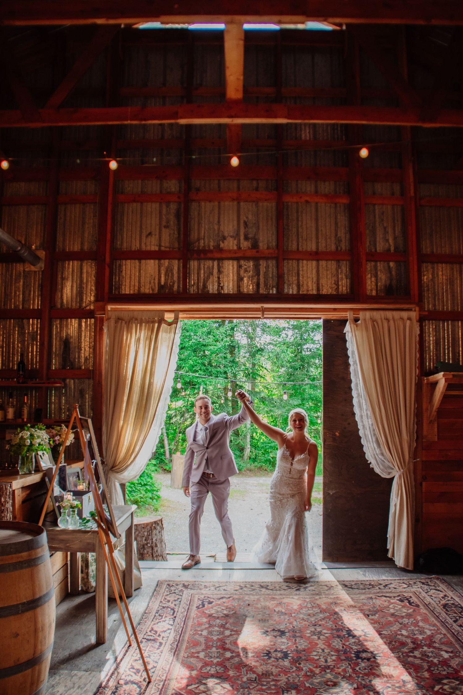 Bride and groom holding hands in rustic barn doorway with lush green trees, string lights, and elegant curtains.