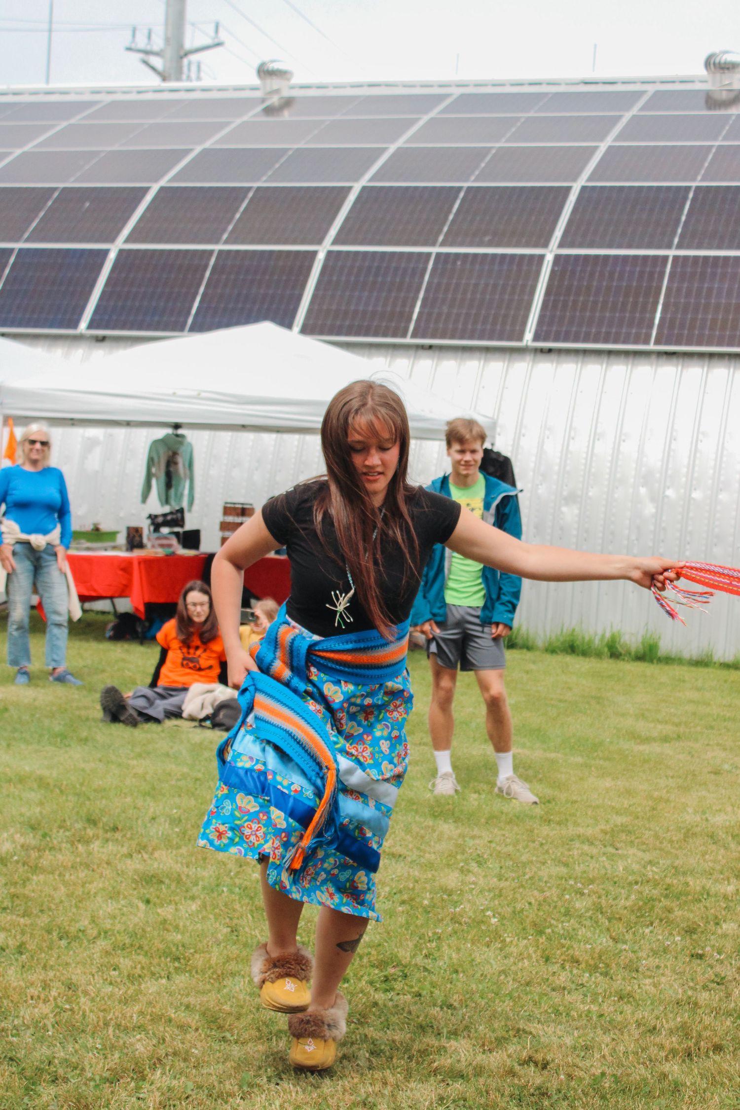 Woman in colorful blue floral skirt and black top dancing outdoors near solar panels at a cultural festival.