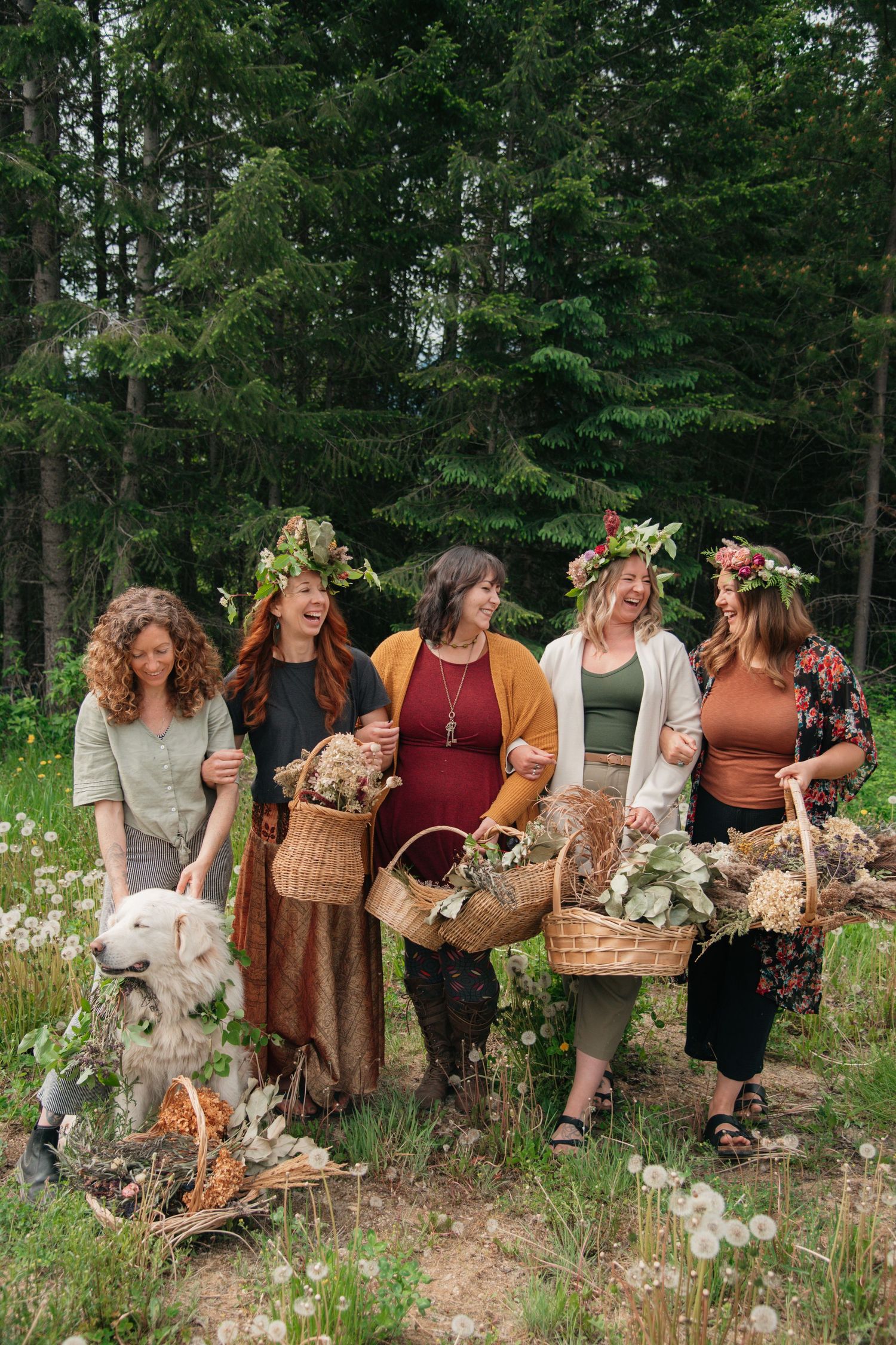 Five women wearing floral crowns holding wicker baskets with wildflowers, standing in a forest meadow with a white dog.