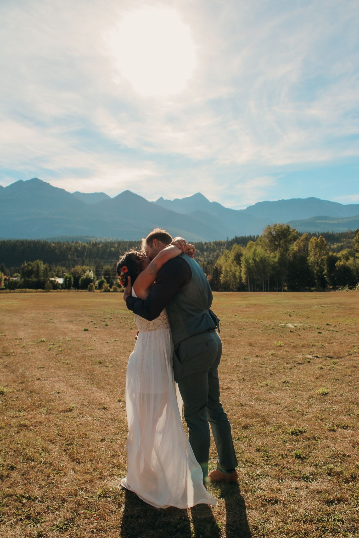 Newlyweds embracing in an open field with dramatic mountain range and forest backdrop.