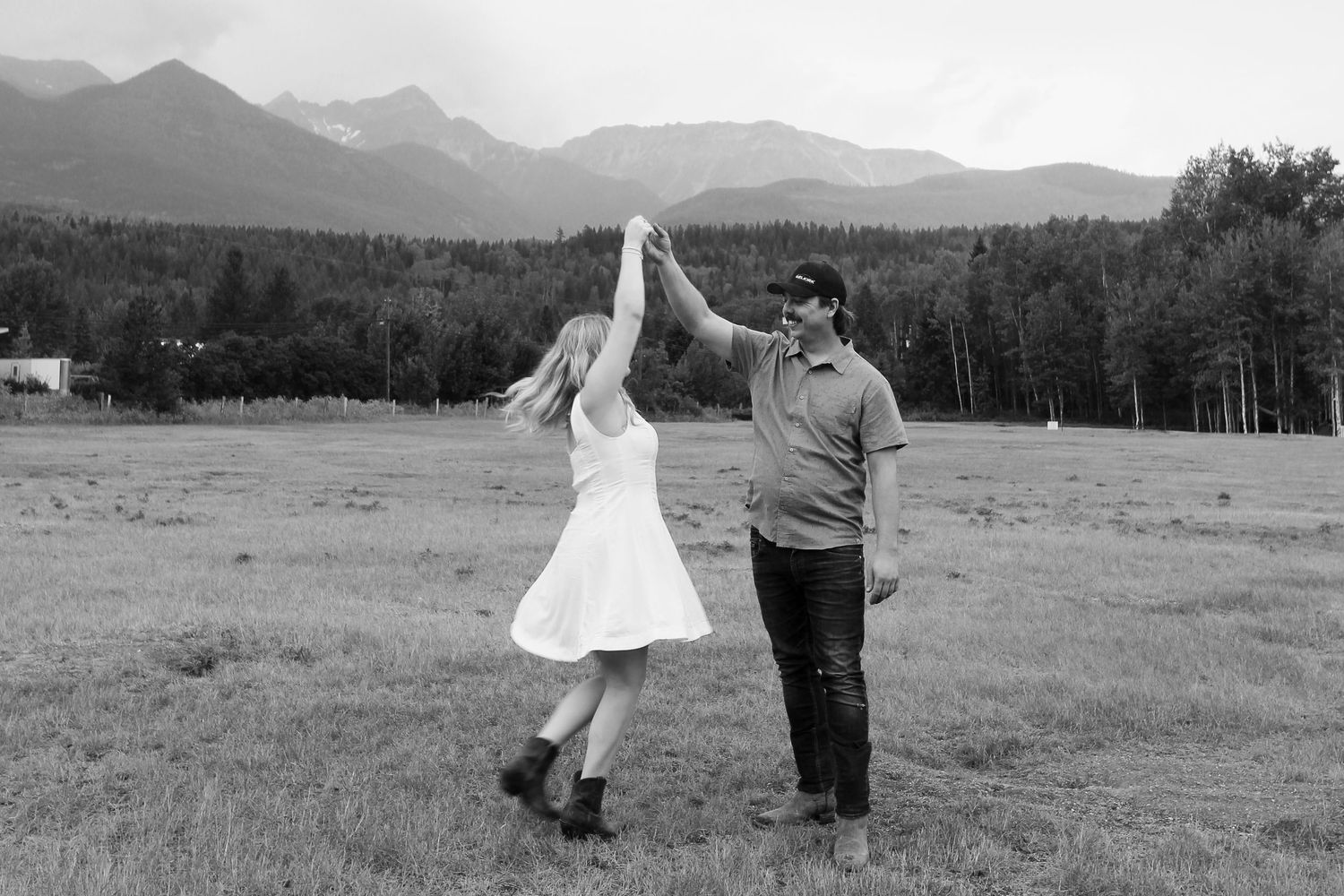 Black and white photo of a couple dancing in a mountain meadow, man spinning woman in white dress.