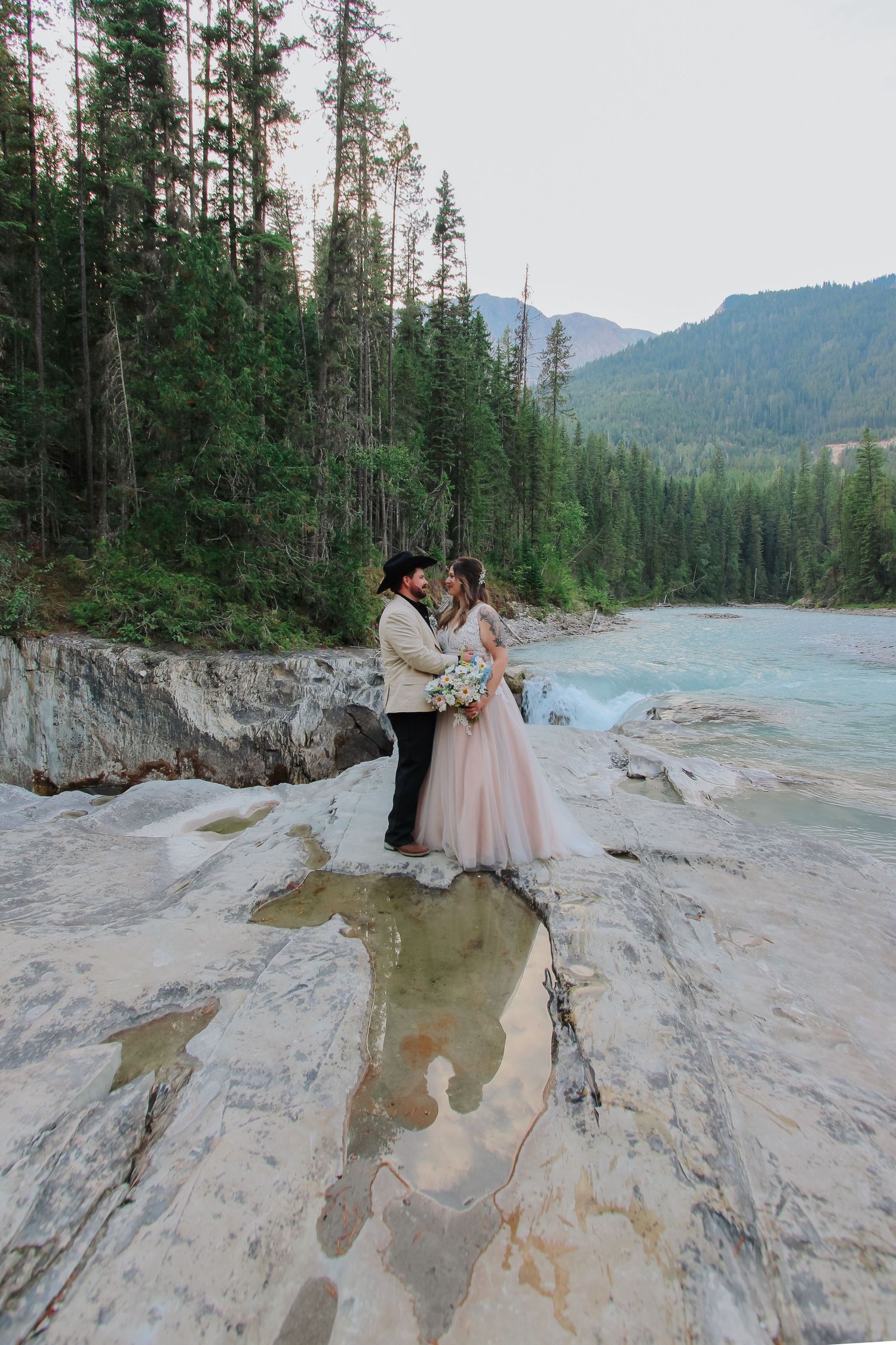 Bride in pink gown and groom kiss on rocky riverbank surrounded by evergreen forest and mountains.