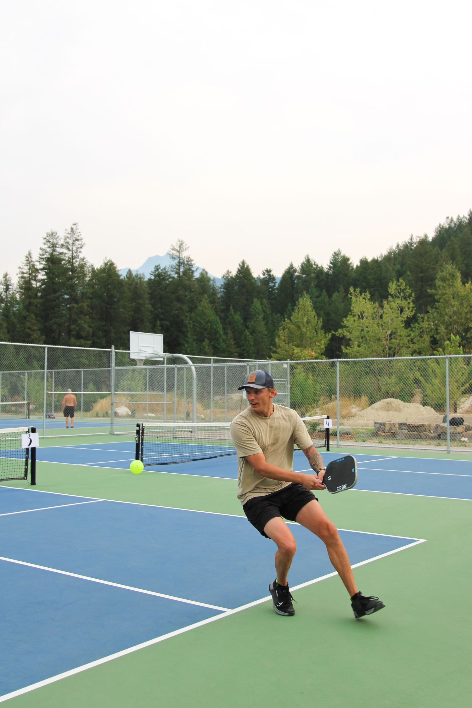 Man playing pickleball on a blue outdoor court surrounded by evergreen trees and mountains in the background.