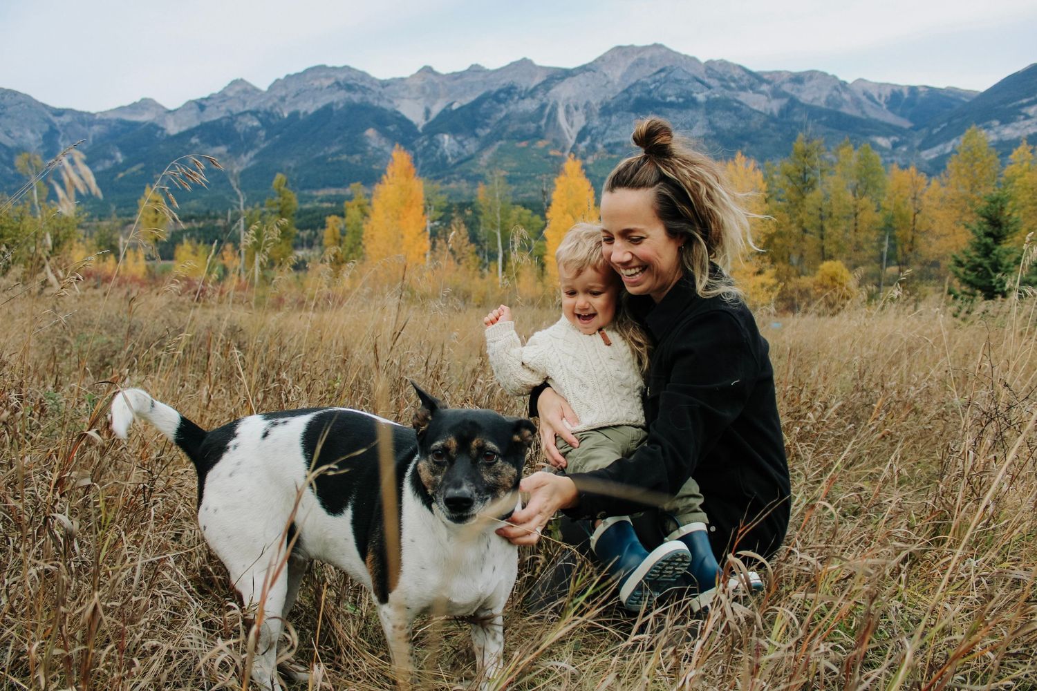 Woman and toddler sitting in autumn field with black and white dog, mountain landscape and golden trees in background.