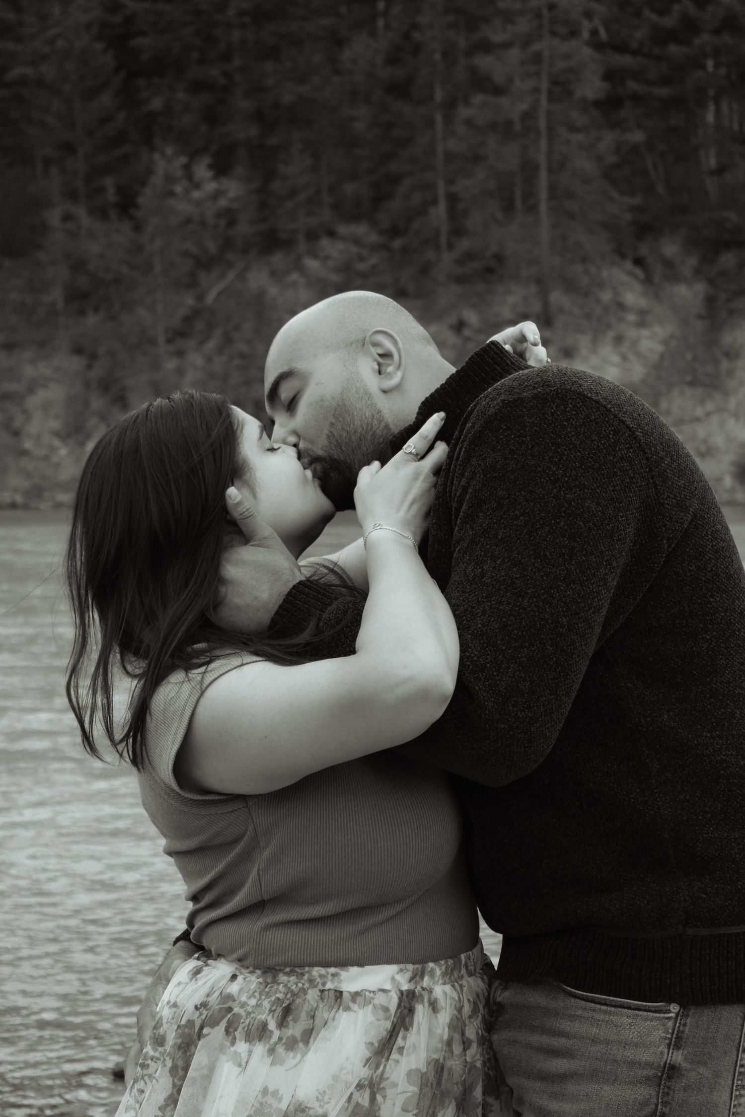 Black and white photo of a couple sharing a romantic kiss near a lake with forest in the background.