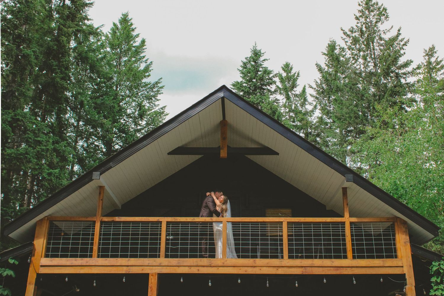 Couple sharing a kiss on the balcony of a rustic wooden cabin surrounded by tall evergreen trees.