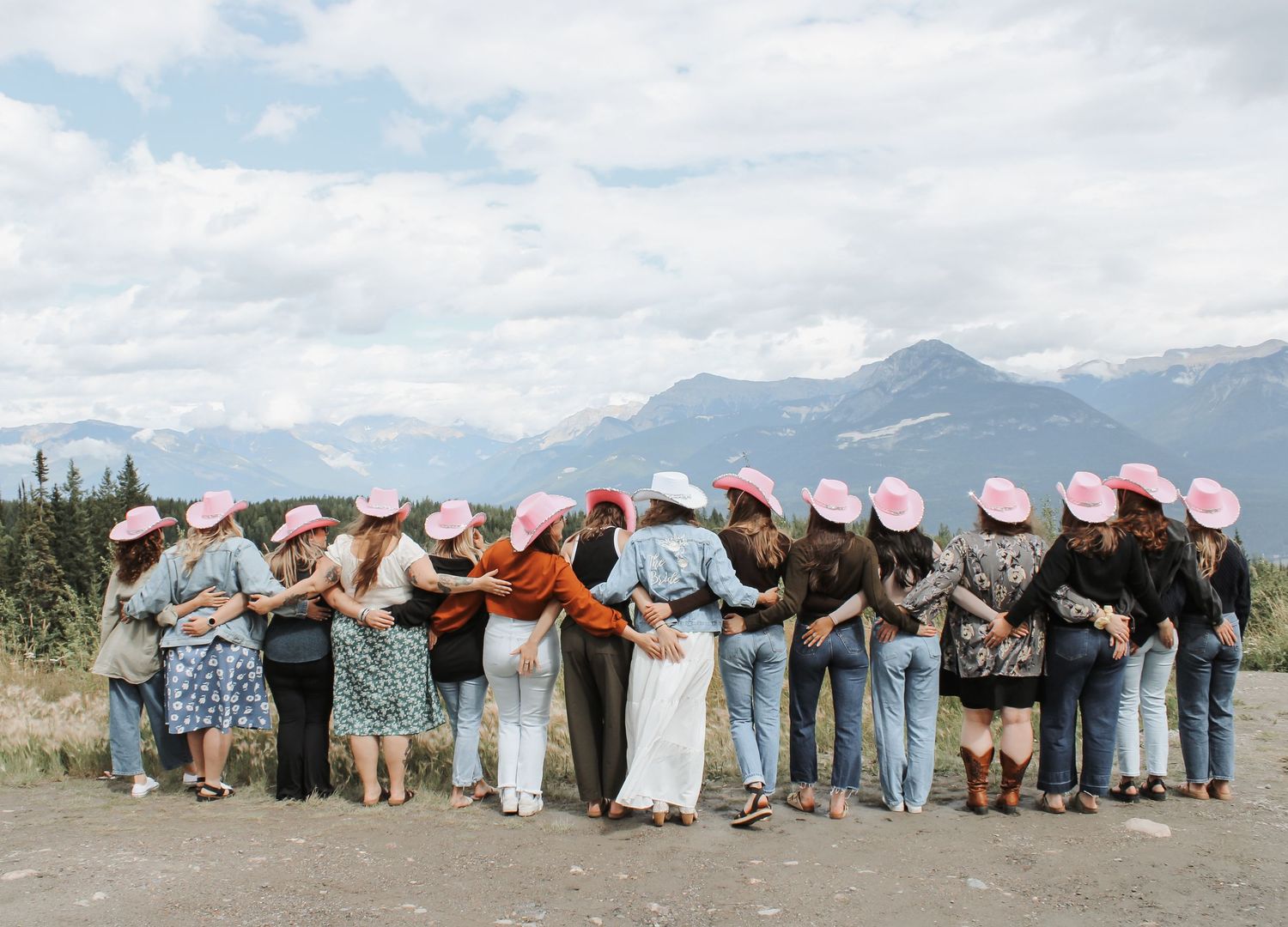 Group of women wearing pink cowboy hats posing with arms around each other against a scenic mountain backdrop.