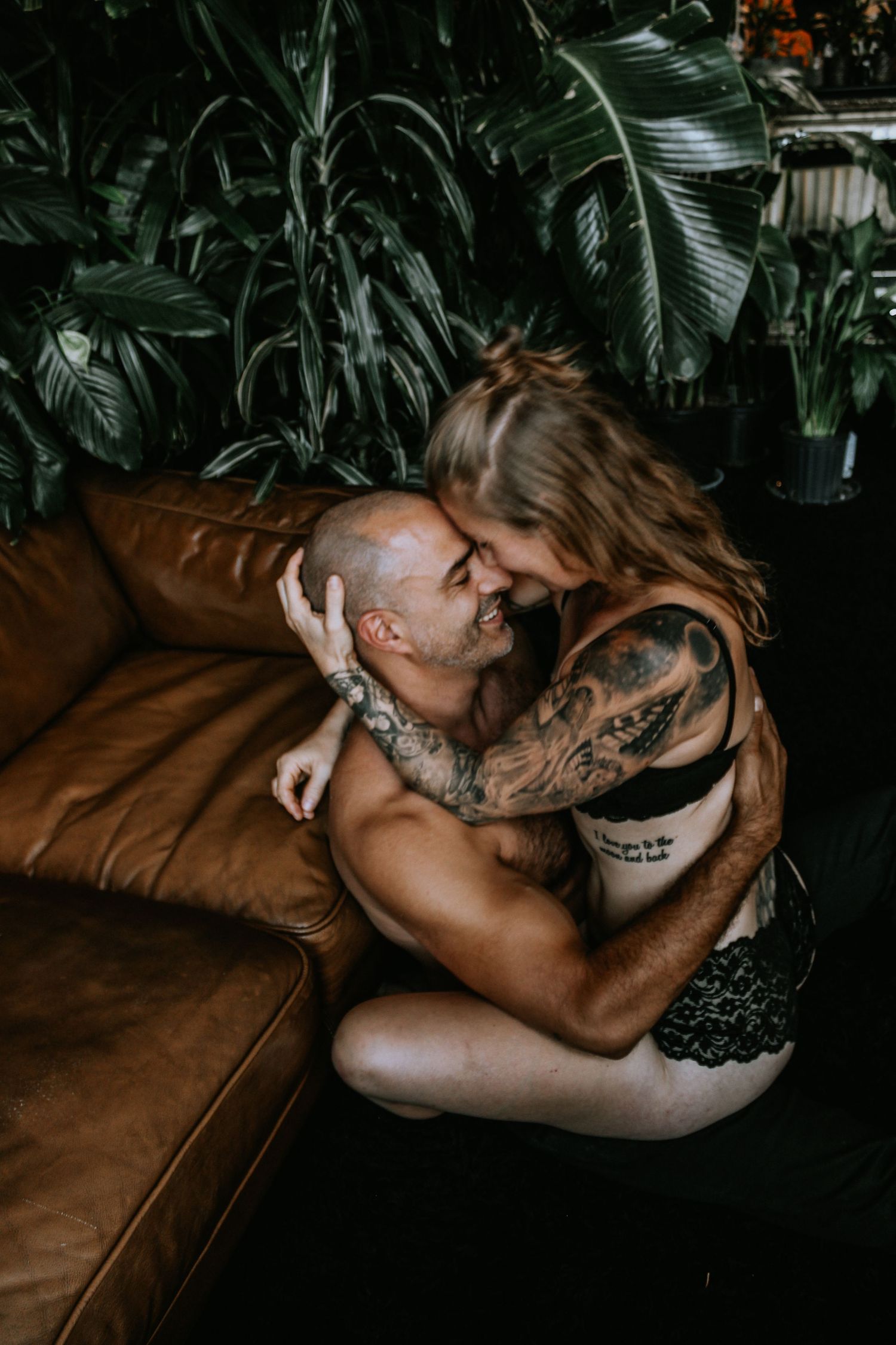 Tattooed couple sharing a romantic embrace on a leather sofa surrounded by lush tropical plants.