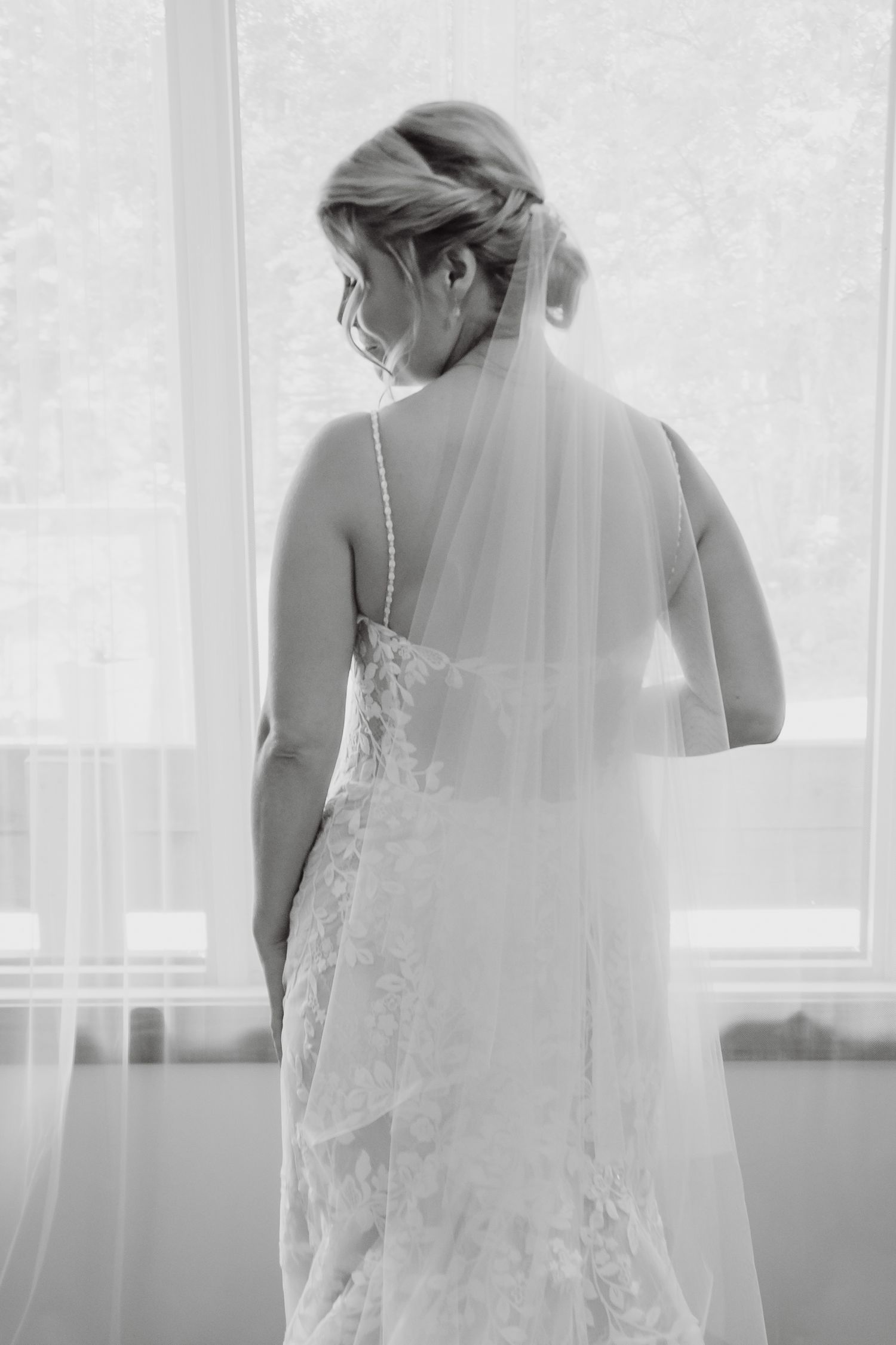 Black and white photo of a bride in a lace wedding gown and veil, standing with her back to the camera near a window.