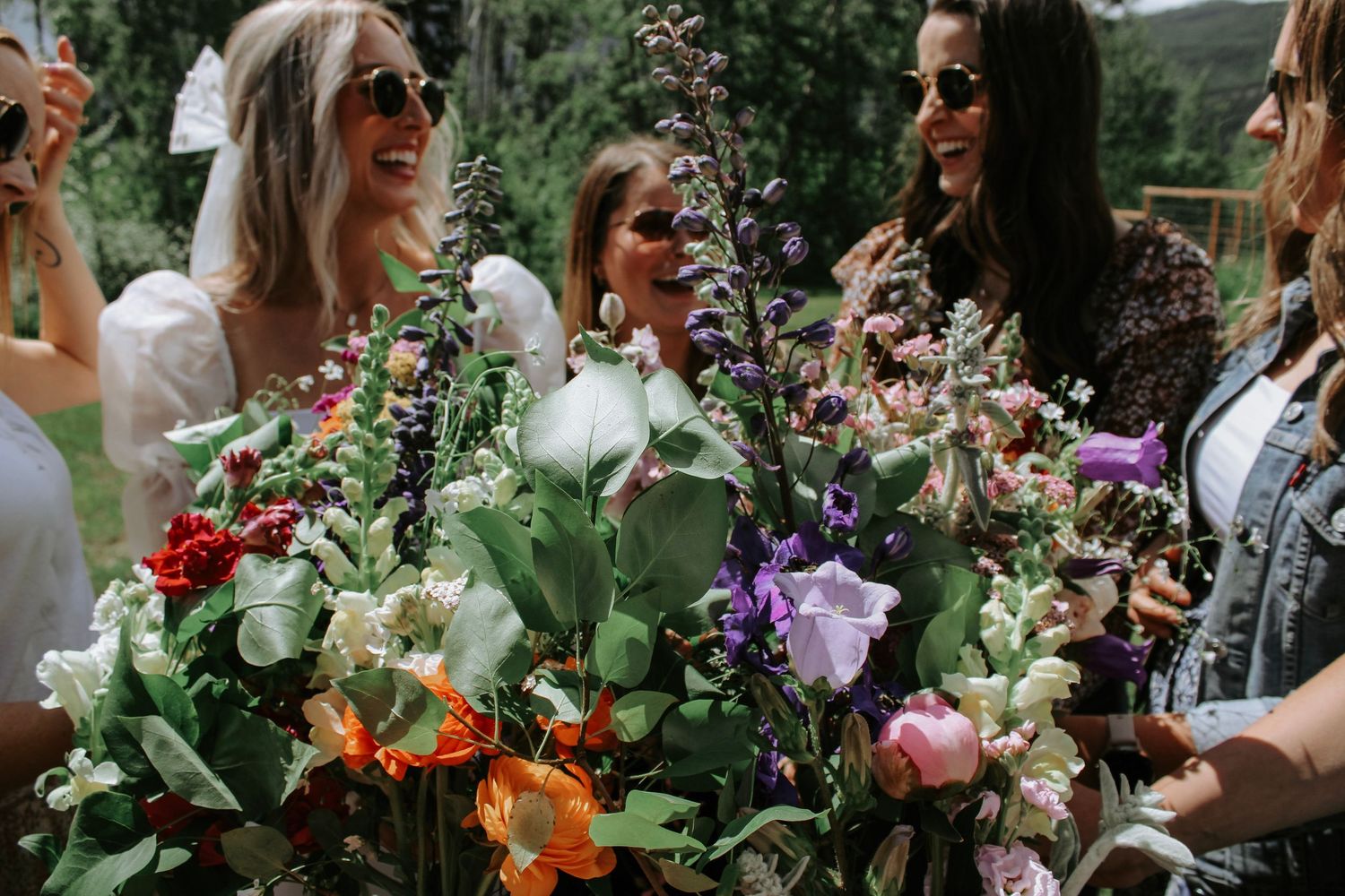 Wedding party members holding vibrant bouquets pose together in a natural outdoor setting.