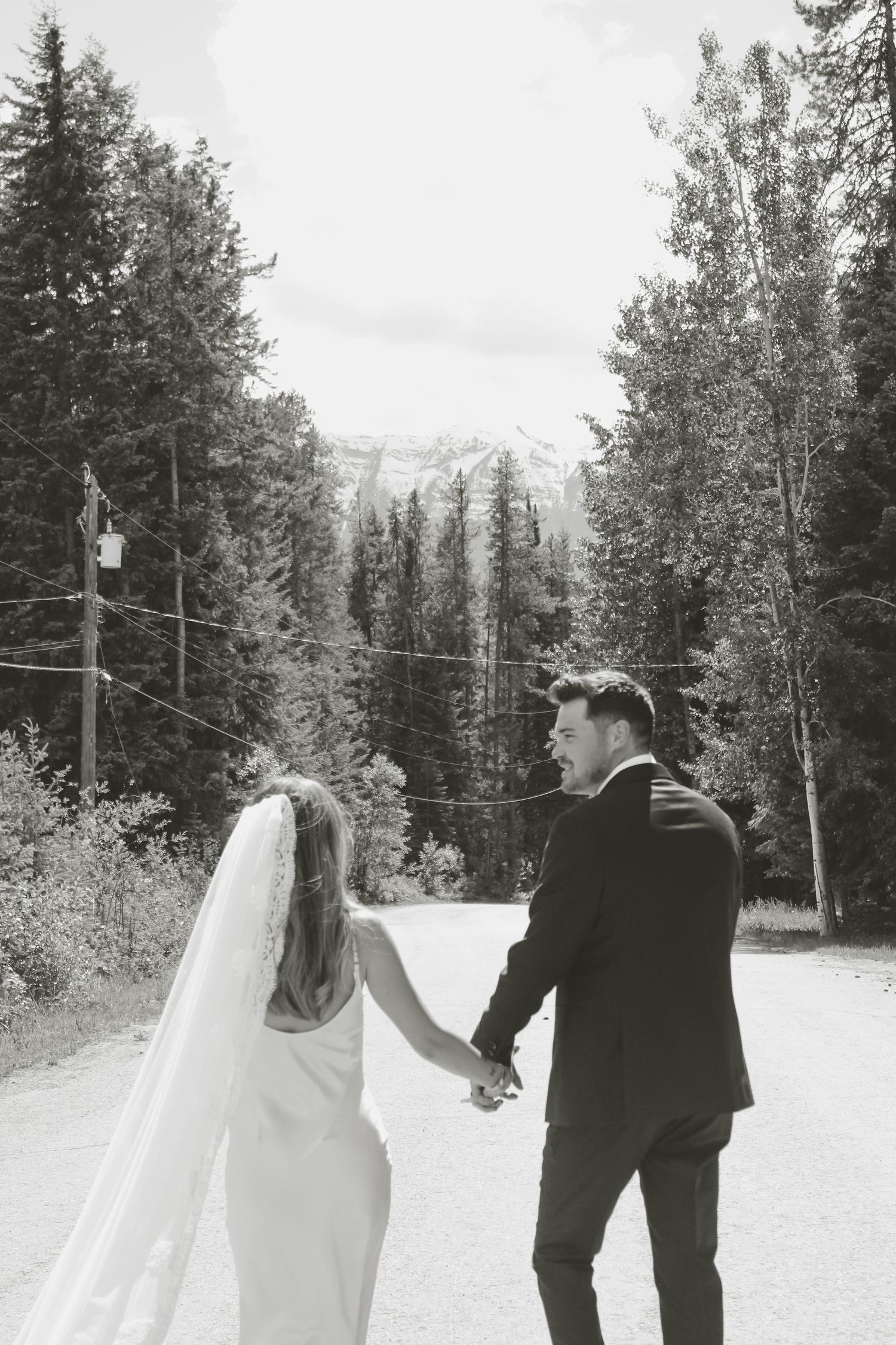 Black and white photo of a bride and groom holding hands walking toward mountain scenery surrounded by evergreen trees.