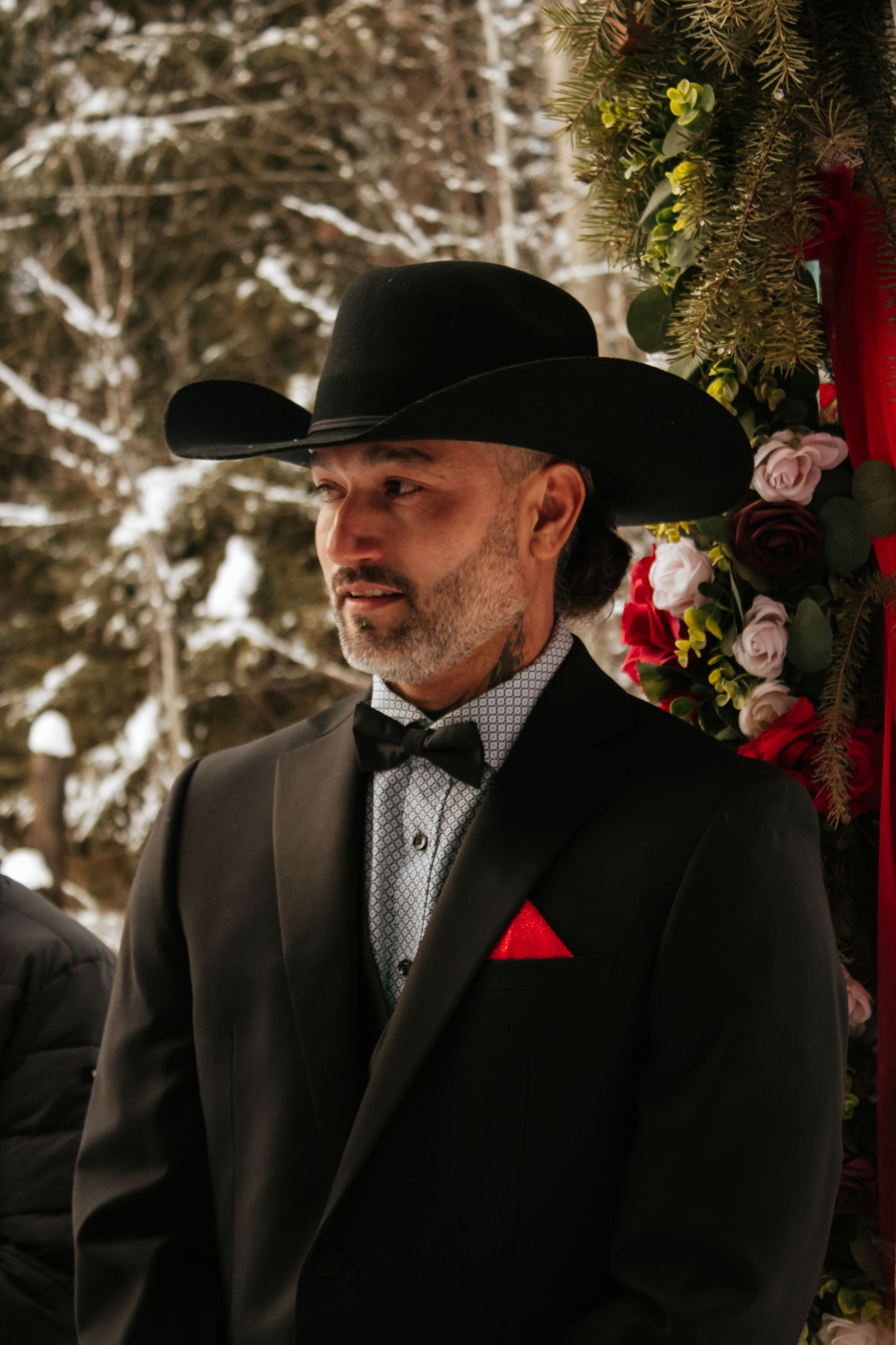 A groom in a black suit and hat stands in front of evergreen branches at a winter wedding ceremony.
