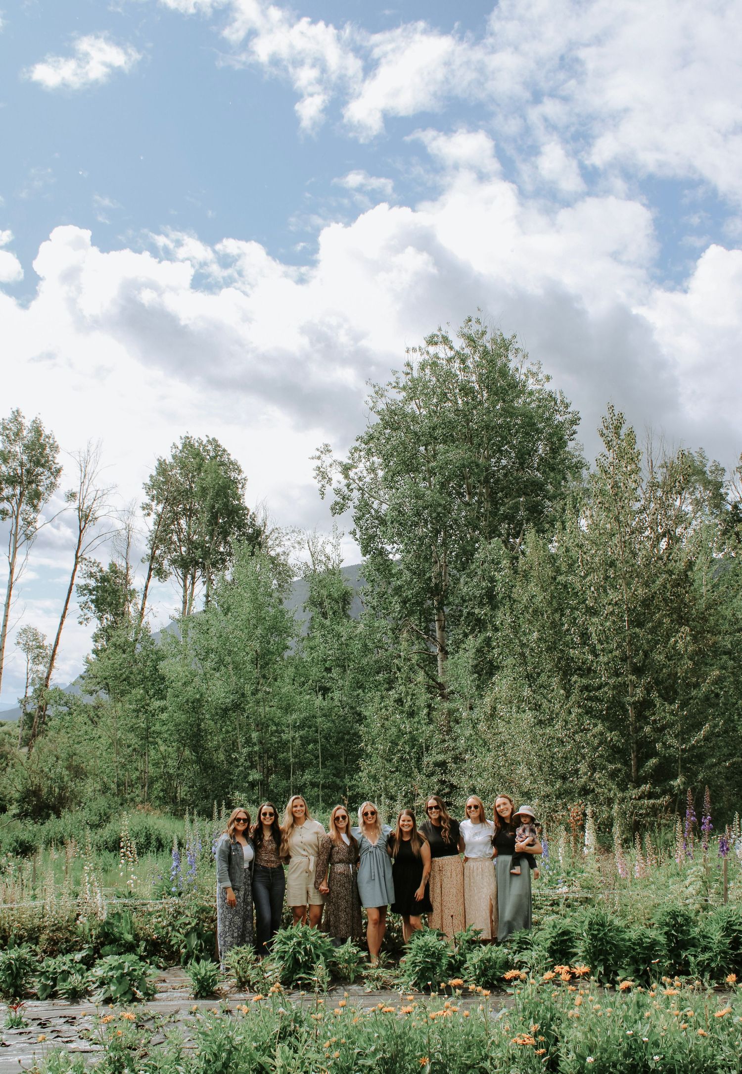 Group of women posing in a lush garden with tall trees and mountains under a cloudy sky.