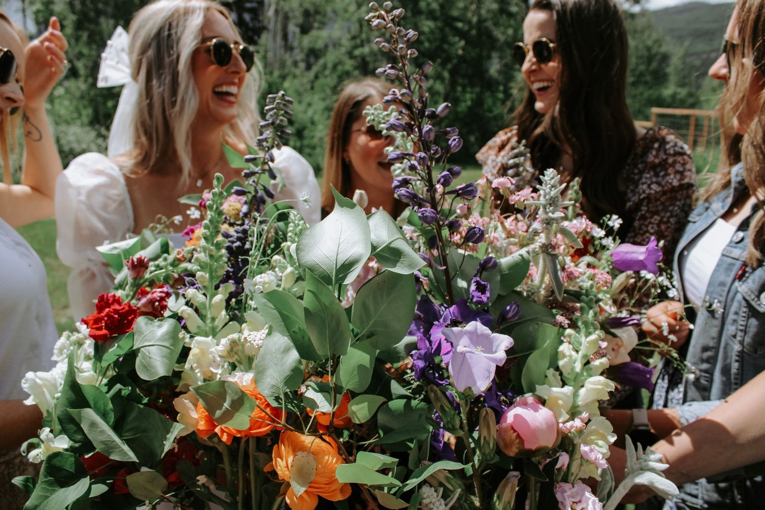 Women laughing and holding colorful wildflower bouquets outdoors at a sunny garden party celebration.