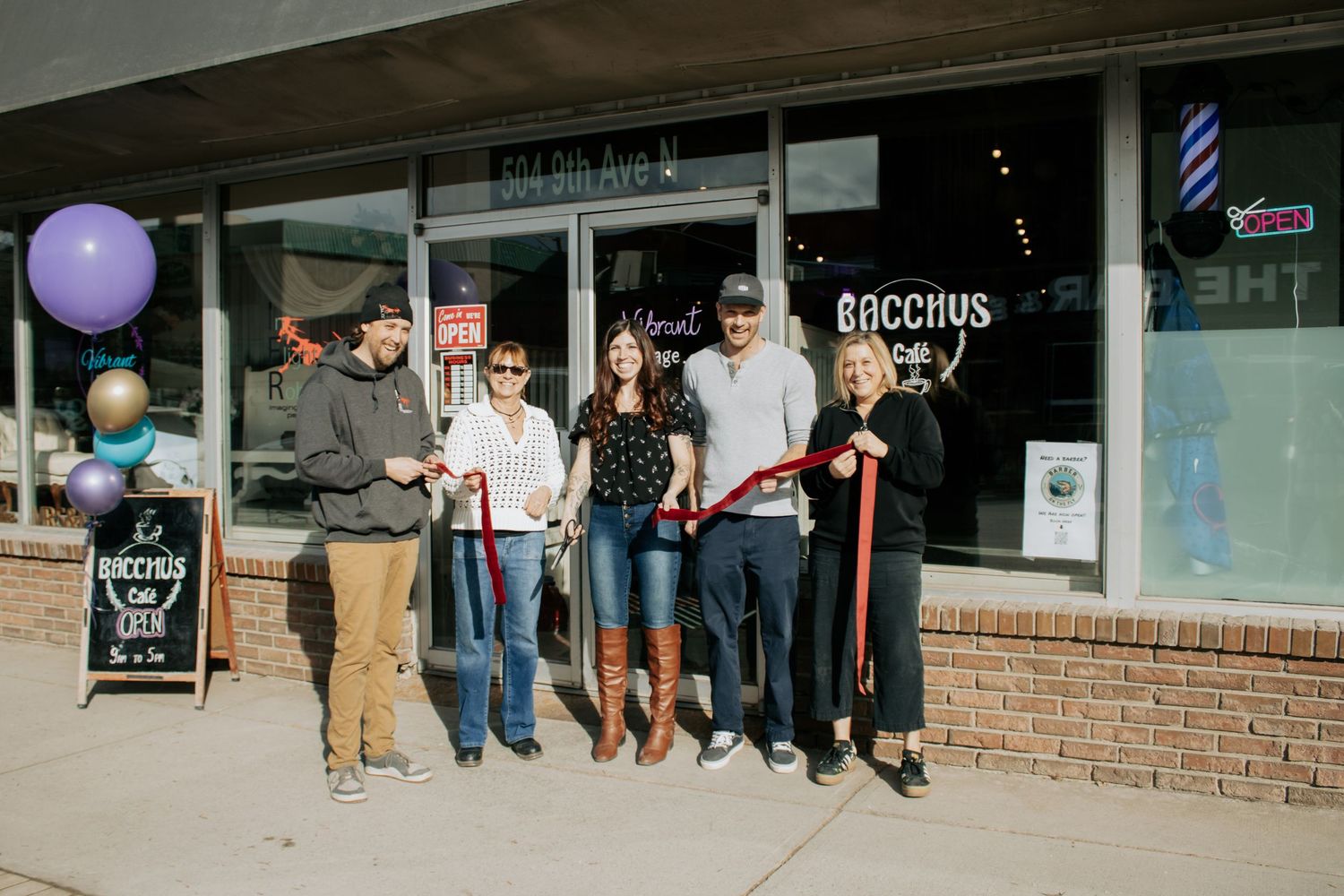 Group of people cutting a ribbon at the grand opening of Bacchus bar on 5th Avenue, with purple balloons outside.