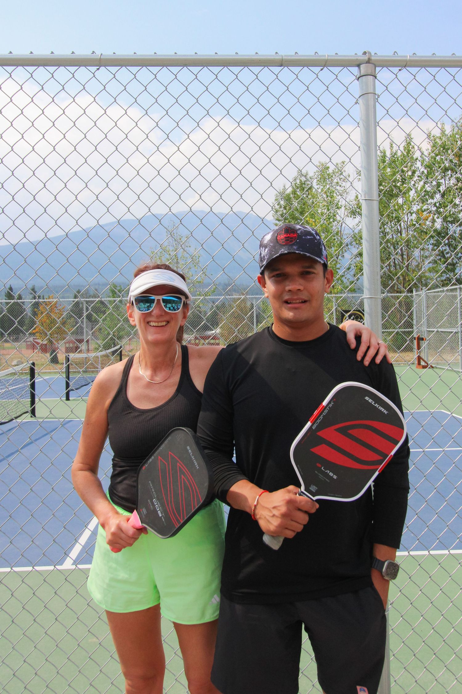 Two pickleball players smile on an outdoor court holding paddles, with mountains and blue sky in the background.
