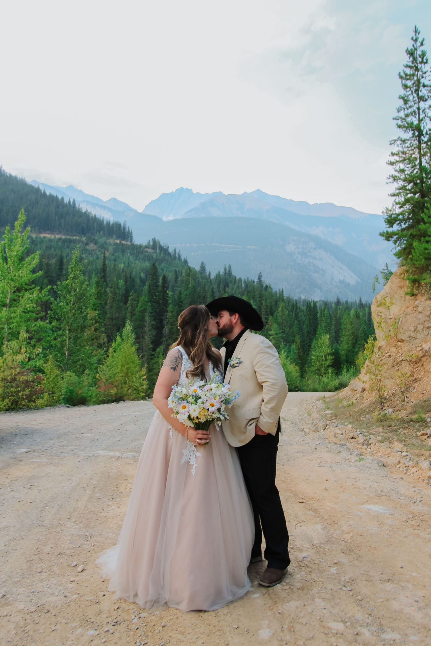 Bride and groom kissing on a mountain road, she holds a wildflower bouquet, peaks behind them.