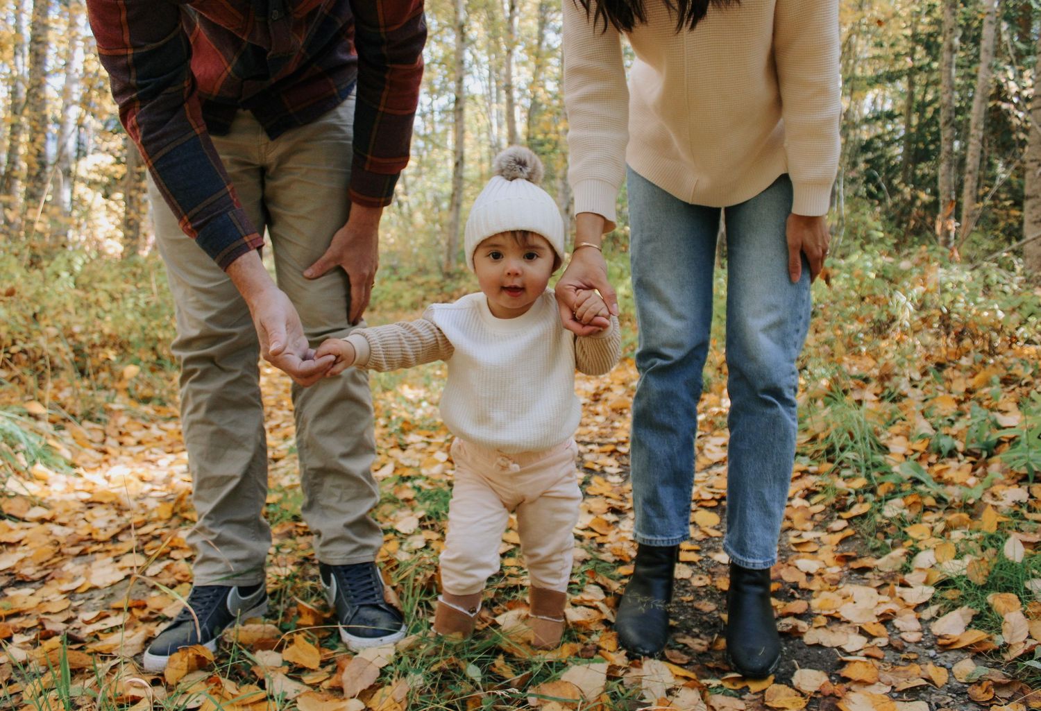 Parents helping their baby take first steps on a leaf-covered forest path in autumn.