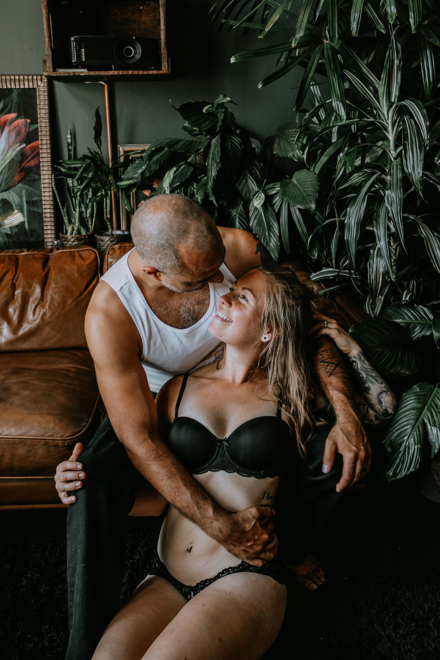 Romantic couple embracing on leather sofa surrounded by indoor plants in moody lighting.