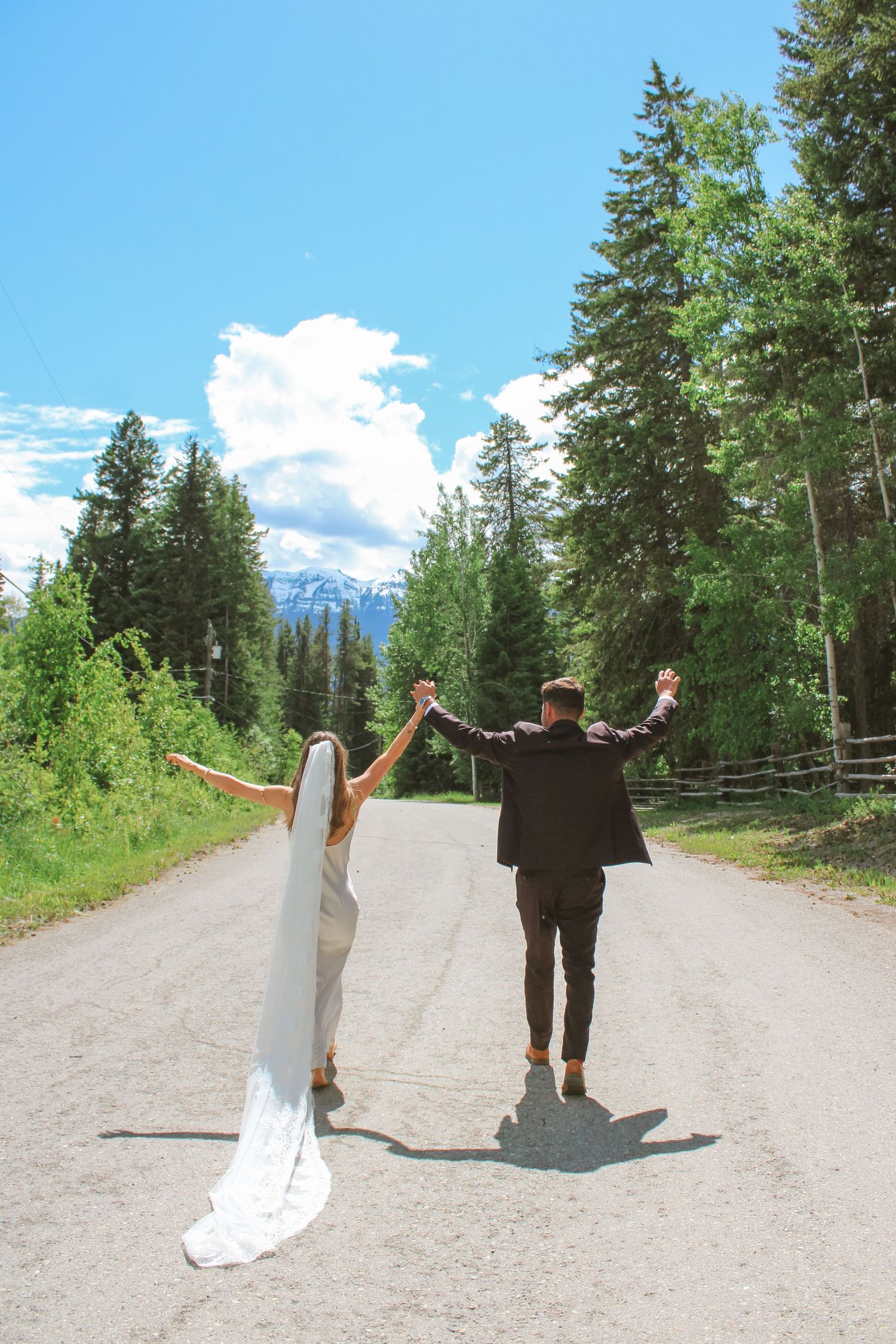 Bride and groom raising hands on mountain road surrounded by pine trees and blue sky on their wedding day.