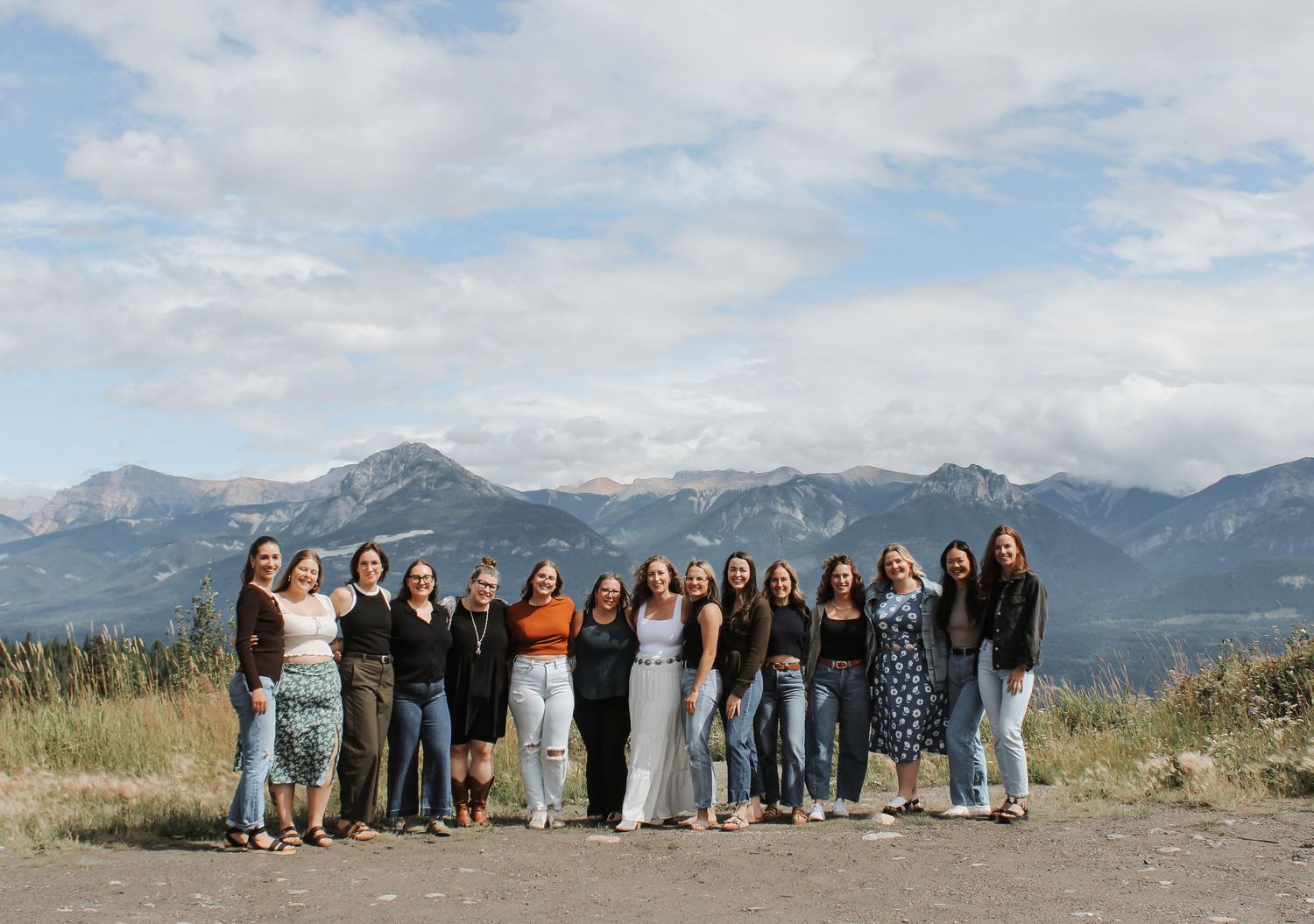 Group of women posing together outdoors with scenic mountain landscape and cloudy sky in background.