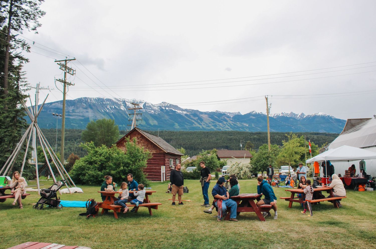 Outdoor gathering with picnic tables on green lawn, log cabin, teepee, and snow-capped mountains in background.