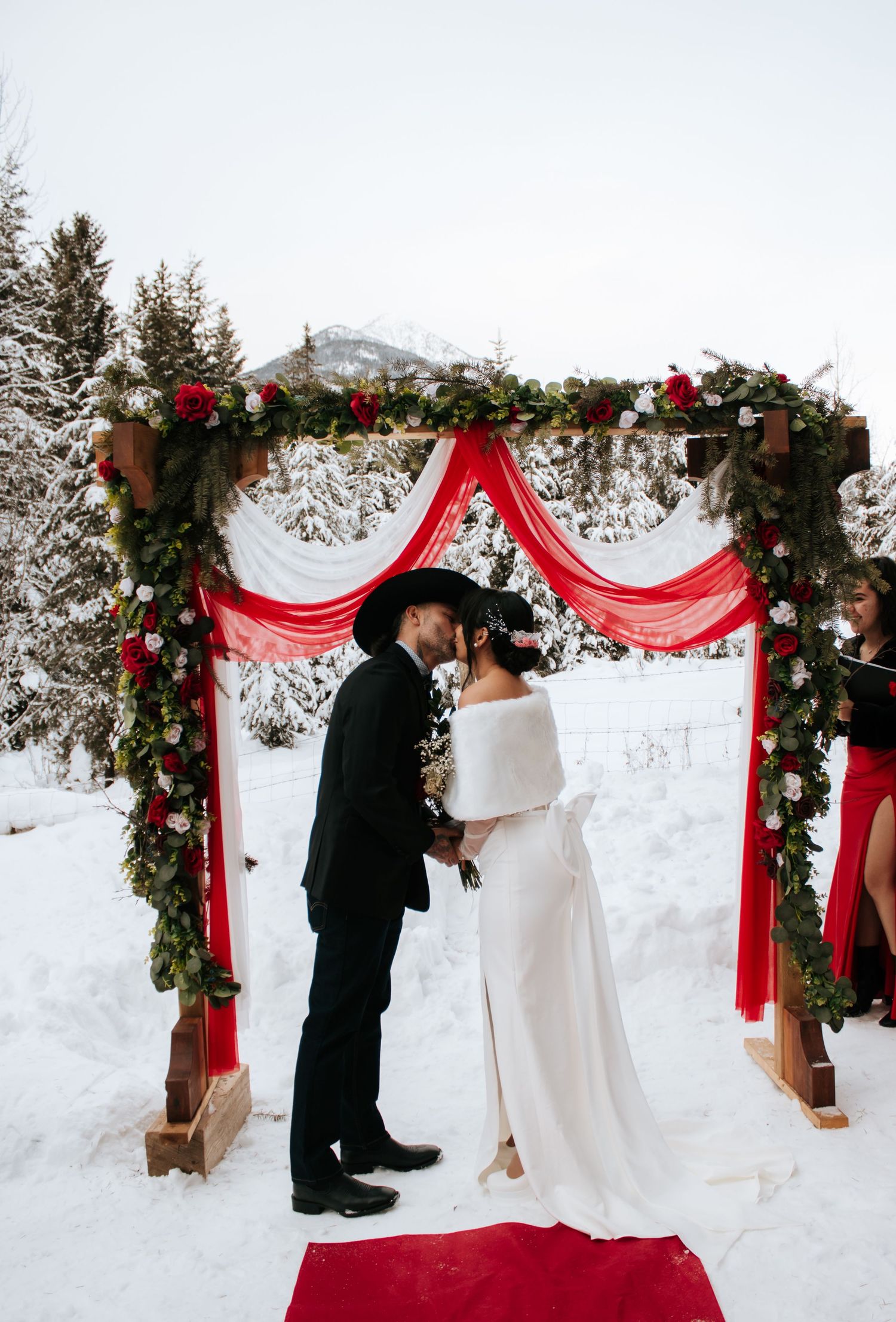 A winter wedding ceremony setup with a red-draped arch, evergreen garlands, and snowy mountain backdrop.