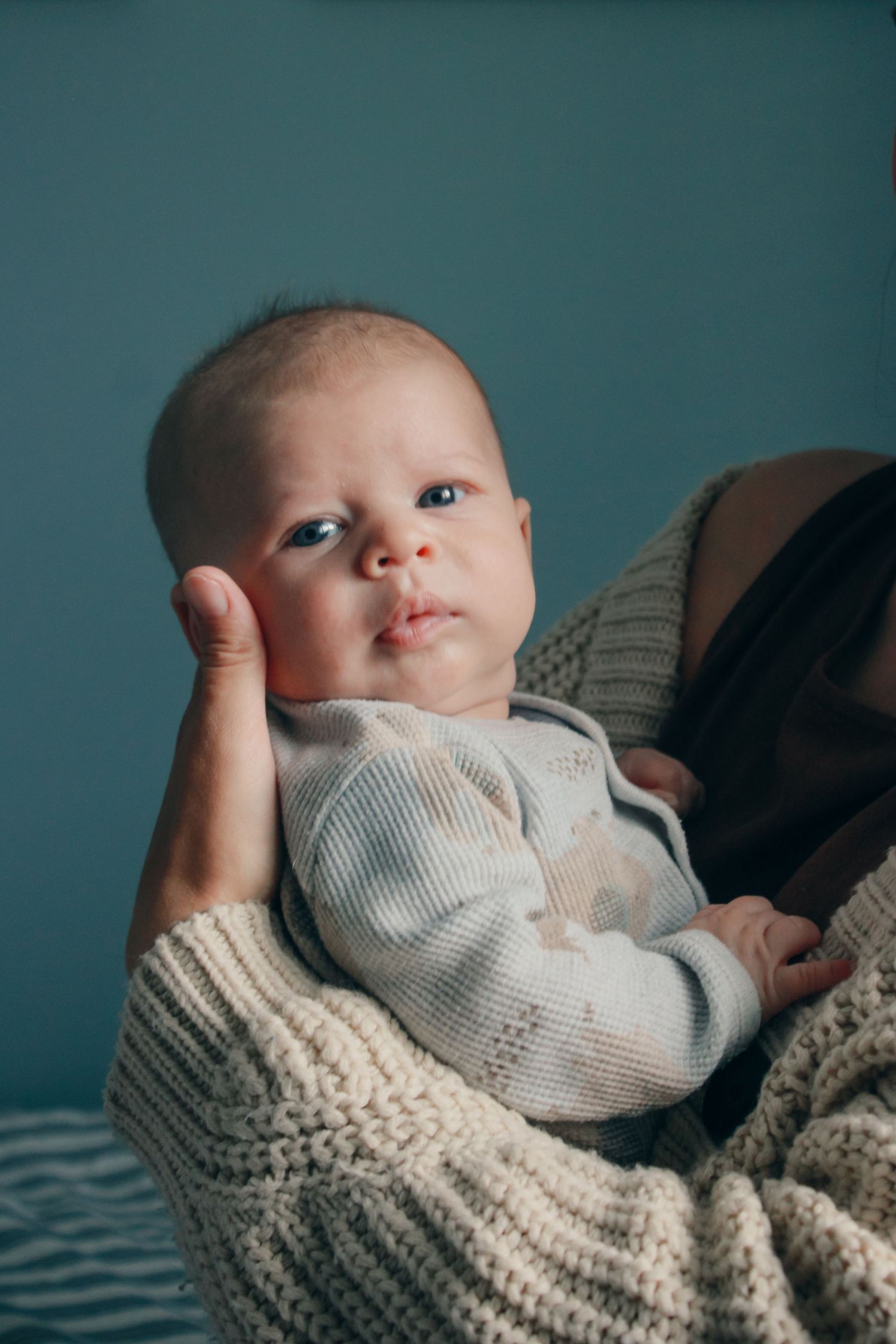 A newborn baby in a striped onesie being cradled in loving arms wrapped in a cozy knit blanket against a teal background.