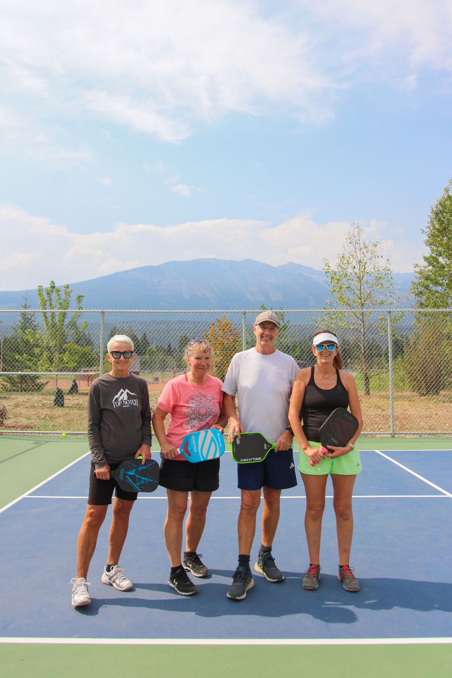Four pickleball players posing on an outdoor court with mountain views and blue sky backdrop.