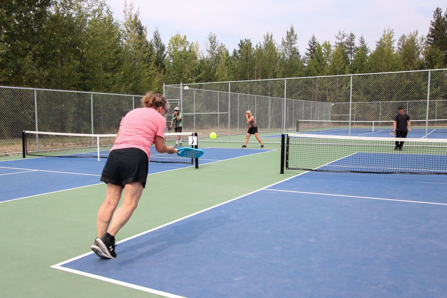 Four players enjoy a pickleball match on a blue outdoor court surrounded by green trees and chain-link fencing.