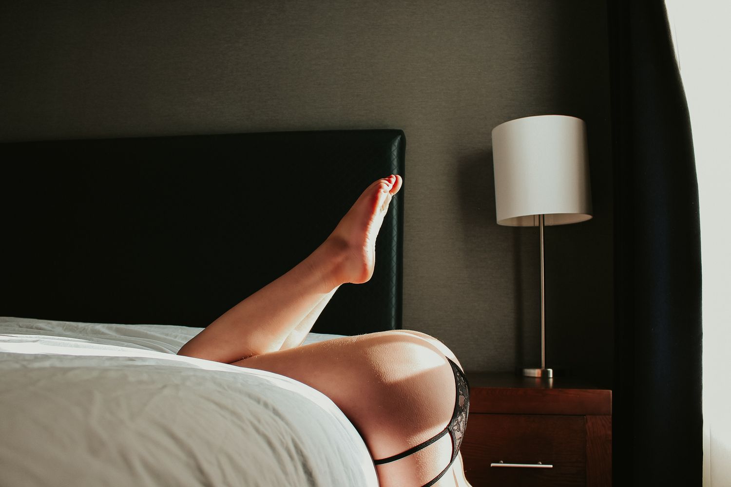Artistic bedroom photo showing legs against dark headboard with bedside lamp.