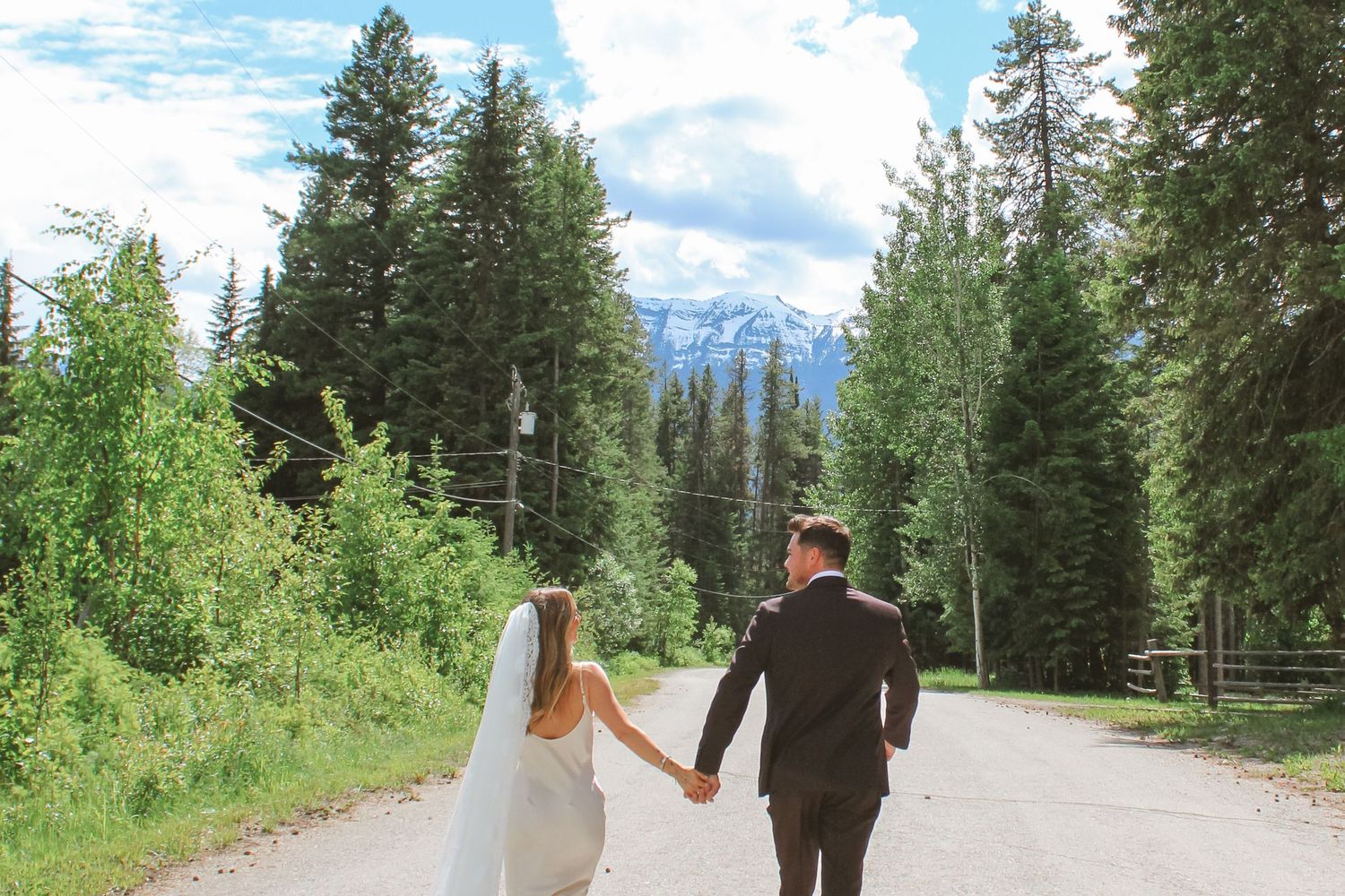 Bride and groom holding hands walking down a scenic mountain road surrounded by tall evergreen trees and snow-capped peaks.