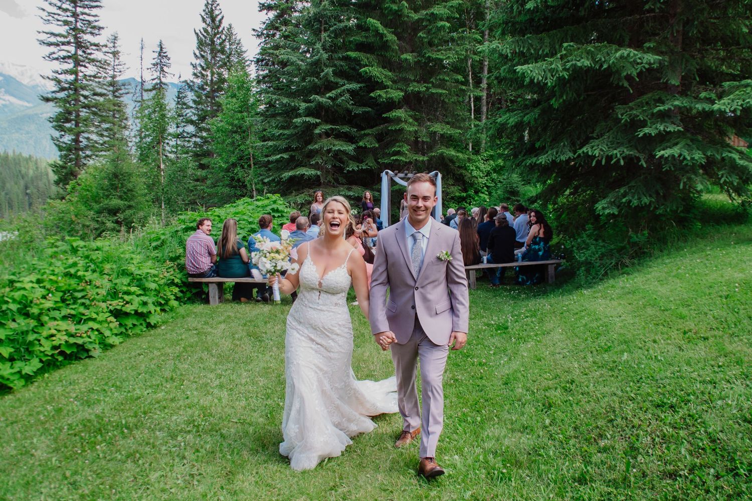 Bride and groom walking down outdoor aisle at mountain wedding ceremony surrounded by tall pine trees.