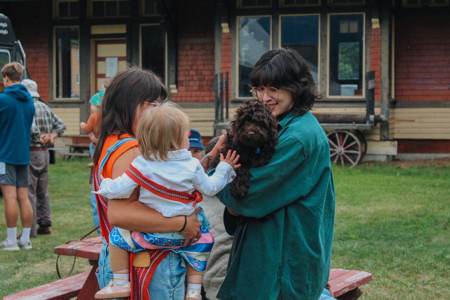 Two women smile outdoors, one holding a baby in a carrier and the other cuddling a small dog near a rustic building.