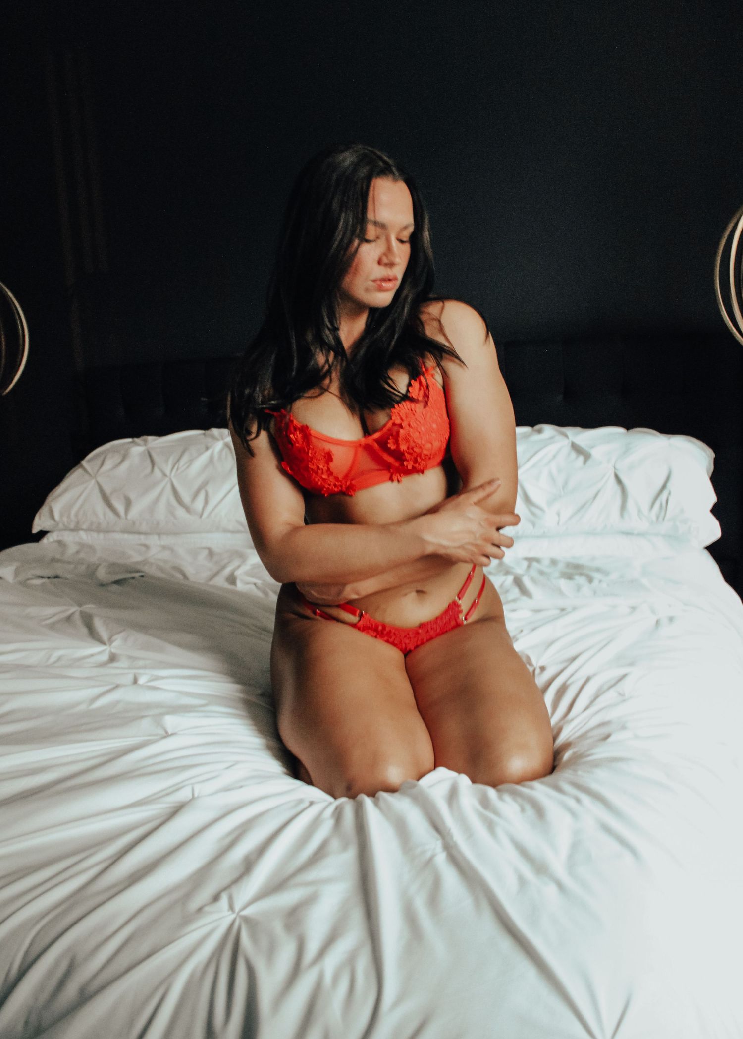 Woman in red lace lingerie kneeling on white bed with arms crossed, dark background.