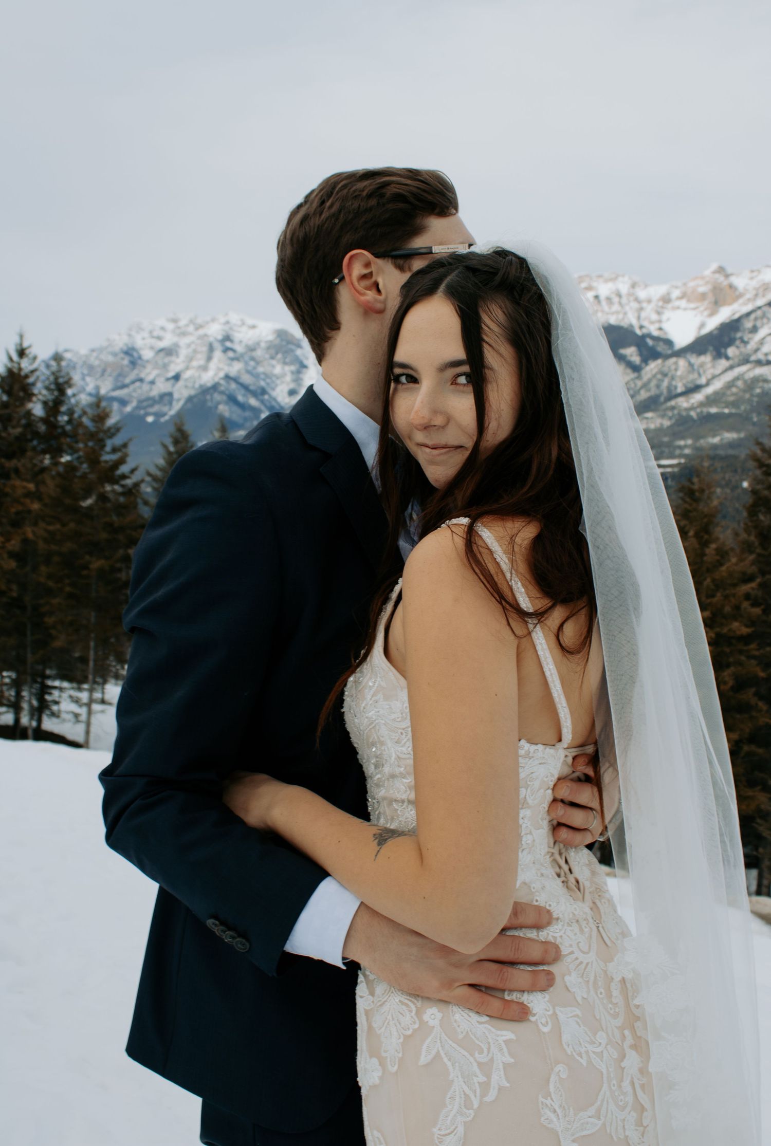 Newlywed couple embraces in a romantic winter mountain setting with snow-capped peaks in the background.