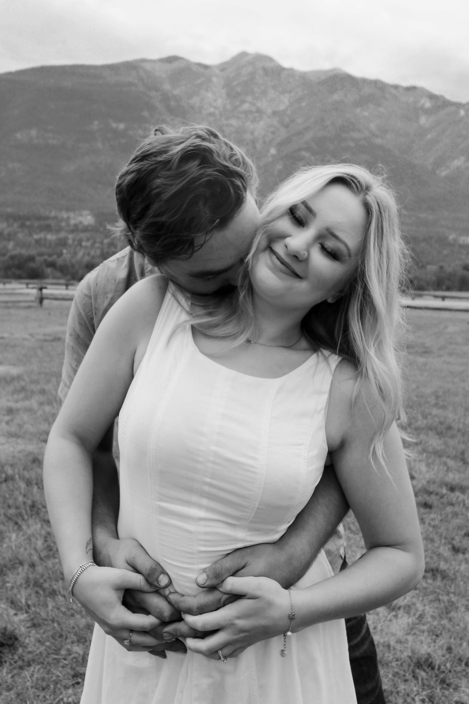 Black and white photo of a man embracing a smiling blonde woman with mountains behind them.
