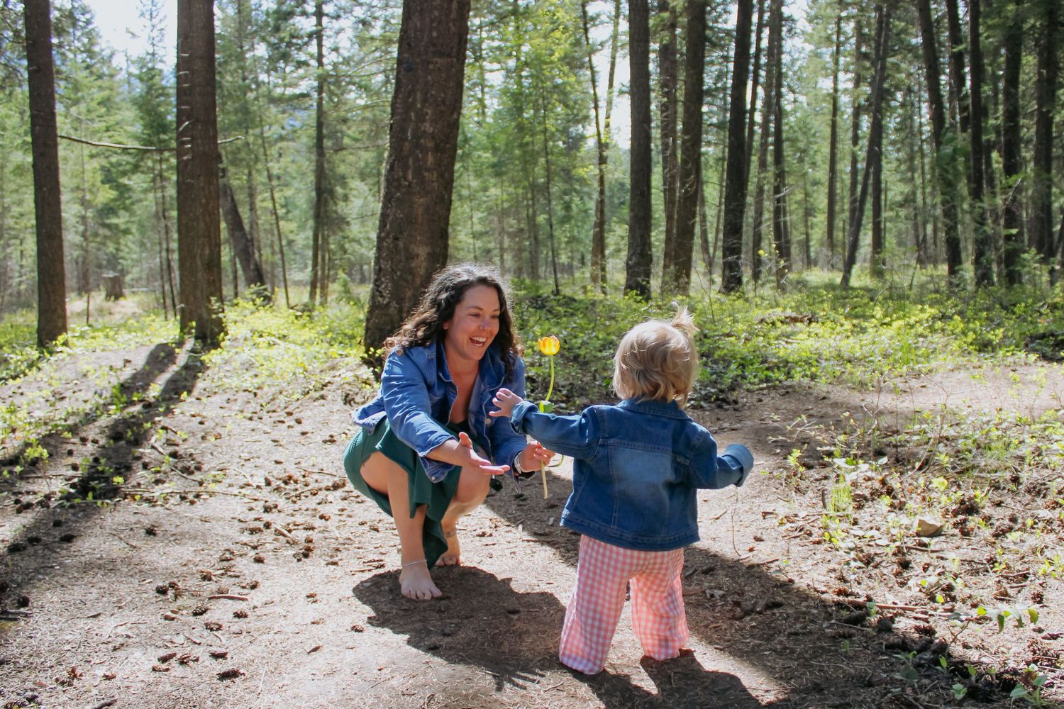 A mother crouches on a forest trail reaching out to her toddler daughter in matching denim jackets among tall pine trees.